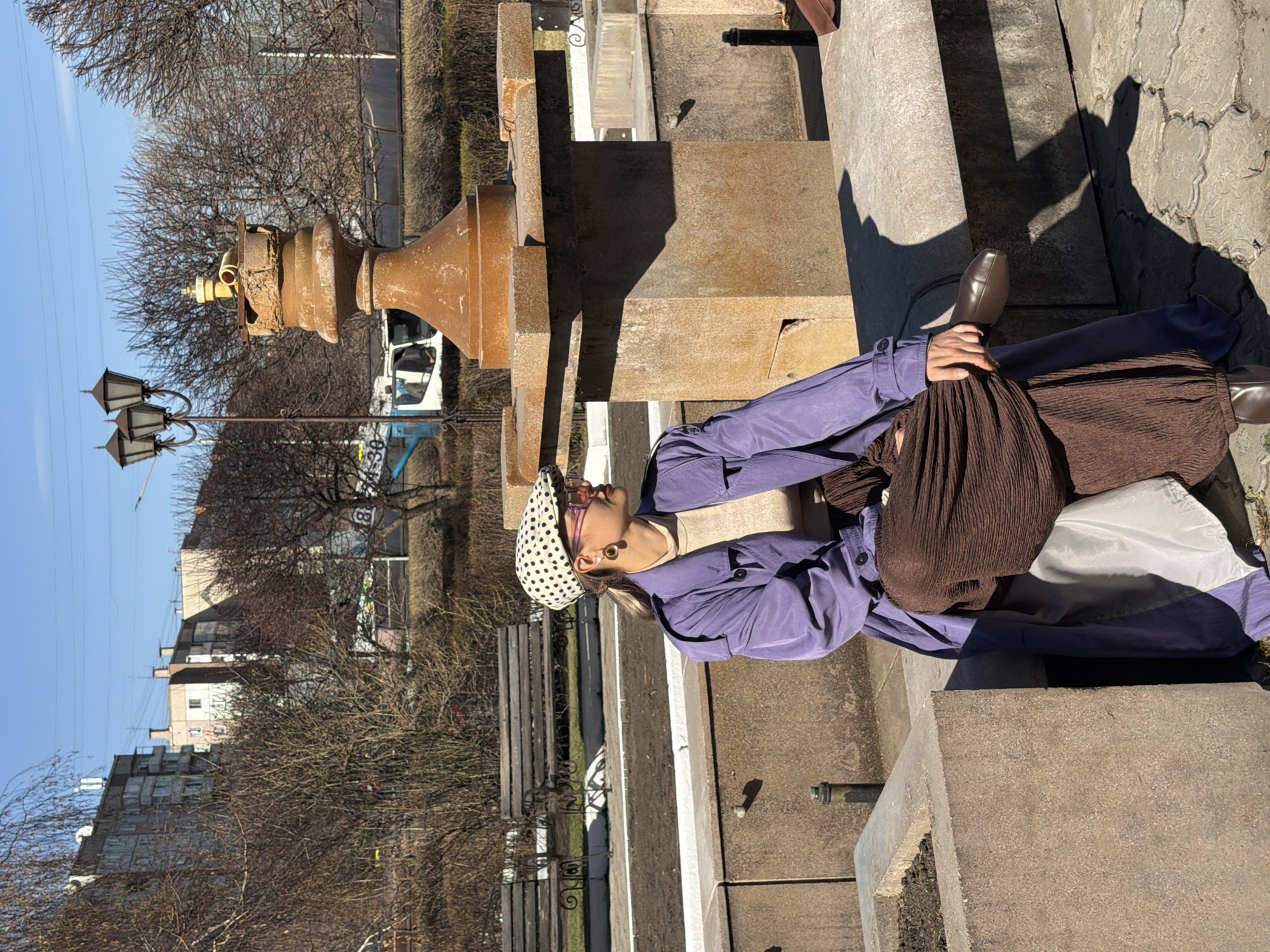 An outdoor scene in Krasnoyarsk, Russia, depicts a person posing on a sunny day, likely in a park or urban square. The subject is a female with blonde hair, wearing a purple long-sleeved top or jacket, a brown pleated skirt, a light beige top, brown shoes, and a white polka-dot beret. She wears glasses and large hoop earrings. She is leaning against a large, weathered stone structure, looking upwards or to the side.
In the foreground and midground, there are multiple worn stone structures, including a large, rusty, ornate metal fixture (possibly part of a decorative fountain or monument) and stone benches. A vintage-style lamppost with three lights stands to the left of the frame. The background features several bare deciduous trees, indicative of winter or early spring. Beyond the trees, multi-story apartment buildings and a white van or small truck are visible.
The bright sunlight casts long, distinct shadows on the ground and stone surfaces, suggesting either early morning or late afternoon. The sky is clear and blue. Visible text includes a white circular sign with a black "B" on a pole in the distance, and partial numbers like "88-38" on the side of the white vehicle.