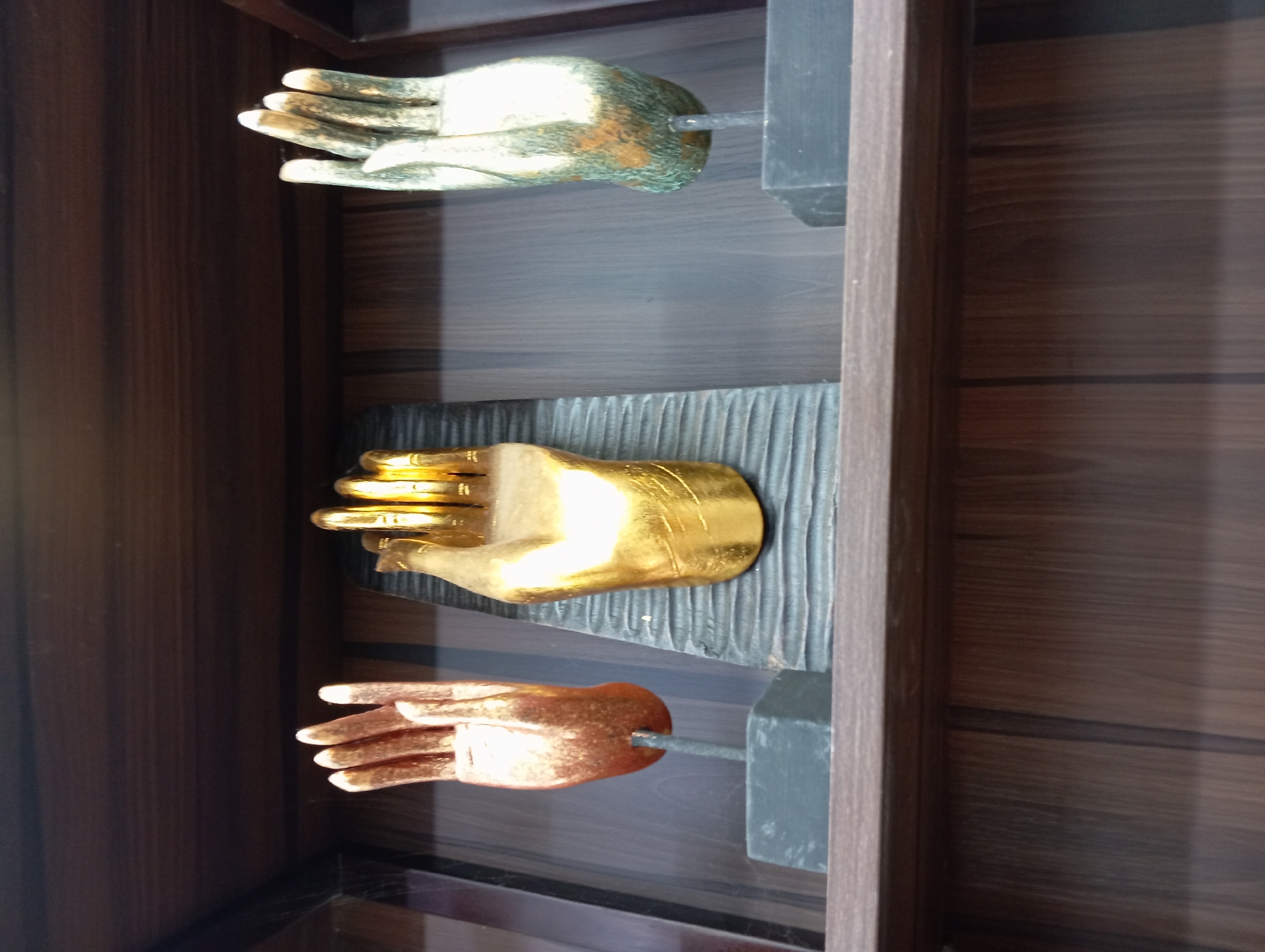 Three decorative hand sculptures are displayed on a wooden shelf. From left to right, the first sculpture is a copper-colored hand with fingers extended, resting on a dark gray square base attached to a thin vertical rod. The middle sculpture is a large, golden hand with all fingers extended, positioned on a dark gray, textured rectangular base. The third sculpture is a metallic hand with a patina of green and gold, also with extended fingers, and mounted on a dark gray rectangular base. The background is a dark wooden paneled wall, and the shelf itself appears to be made of dark wood. The image was taken at the Vinpearl Golf Nha Trang, located in Nha Trang, Vietnam.