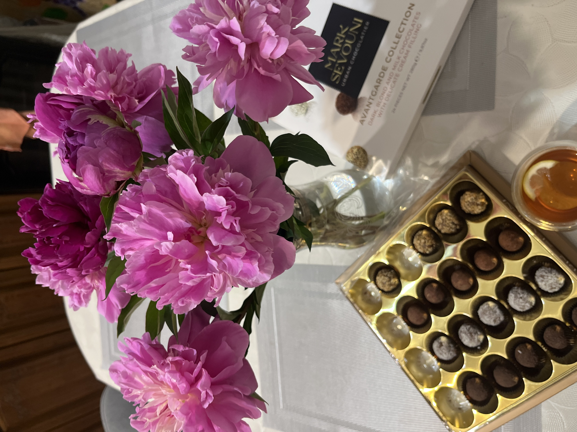 A still life composition captured from an overhead perspective displays a bouquet of vibrant pink peonies arranged in a clear glass vase. Adjacent to the flowers is a white box of "MARK SEVOUNI URBAN CHGEGLATIER AVANTGARDE COLLECTION" chocolates, with text indicating they are dark, blond, and milk chocolates with delicate cream filling. The box contains 24 pieces and weighs 200g. An opened gold box of similar chocolates reveals an assortment with various coatings. A small cup of tea with a lemon slice sits near the chocolates. The setting is a surface covered with a white tablecloth. The location is not discernible from the image.