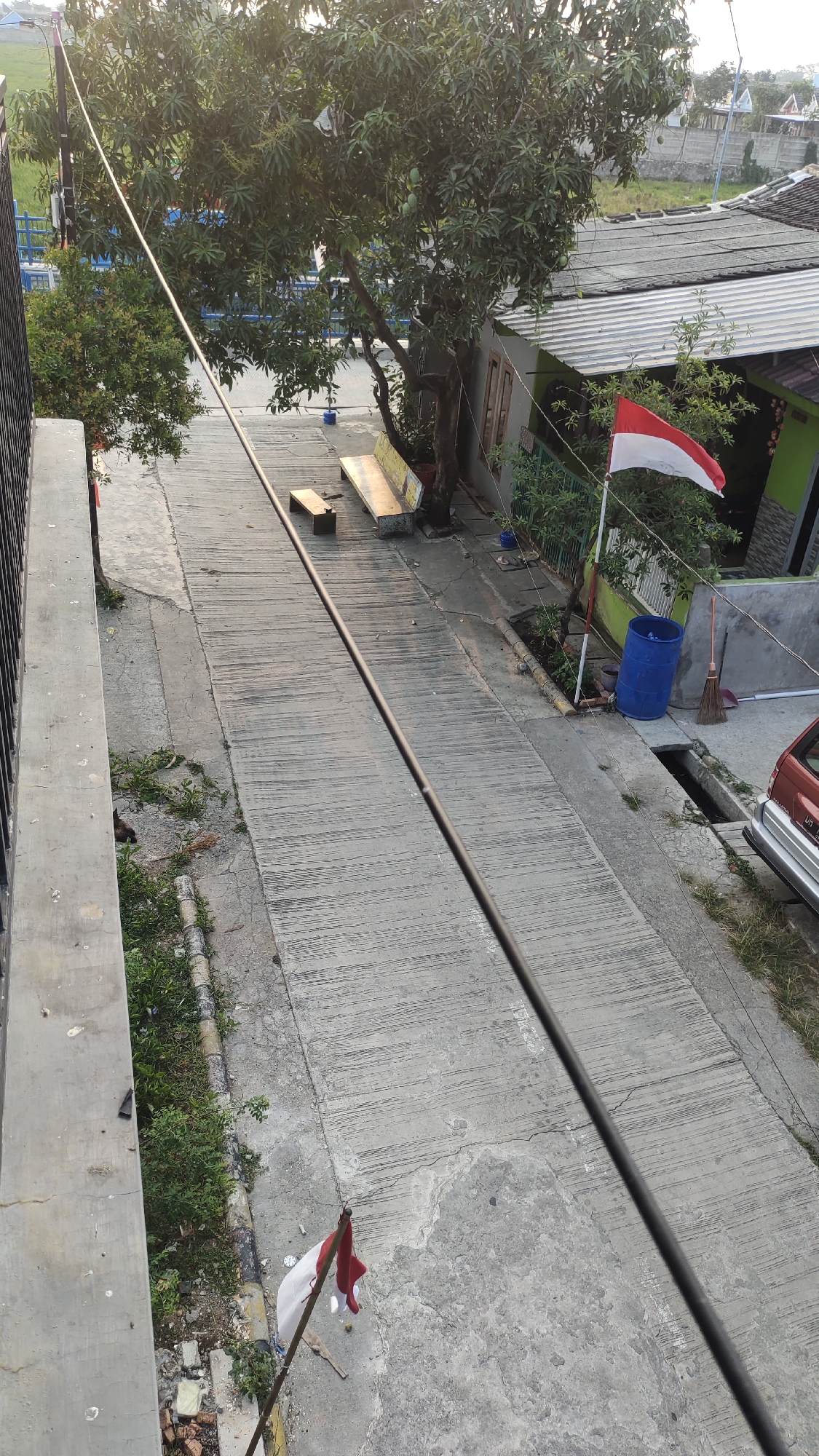 The image shows an outdoor scene in Jalenjaya, Indonesia, under natural daylight. In the foreground, a concrete walkway or road occupies a significant portion of the frame, with a thin dark cable running diagonally across it. To the left, a concrete wall borders the walkway, with some greenery and debris at its base. Further down the walkway, a small Indonesian flag, red and white, is planted on a thin pole.
In the midground, a large tree with dense foliage stands prominently. Beneath the tree, there are two benches. One is a long, slatted yellow bench, and beside it is a smaller, wooden bench. Behind the tree, a building with a grey roof and green walls is visible. An Indonesian flag, also red and white, is mounted on a pole in front of this building. To the right of the building, a blue plastic barrel and a broom are situated near a drainage ditch. A portion of a red car is visible parked near the drainage.
In the background, houses with grey roofs and a light-colored wall are visible, suggesting a residential area. The sky is visible at the top, appearing bright and clear. There are no people clearly depicted in the image. The overall environment appears to be a street or pathway in a village or suburban setting within Jalenjaya.