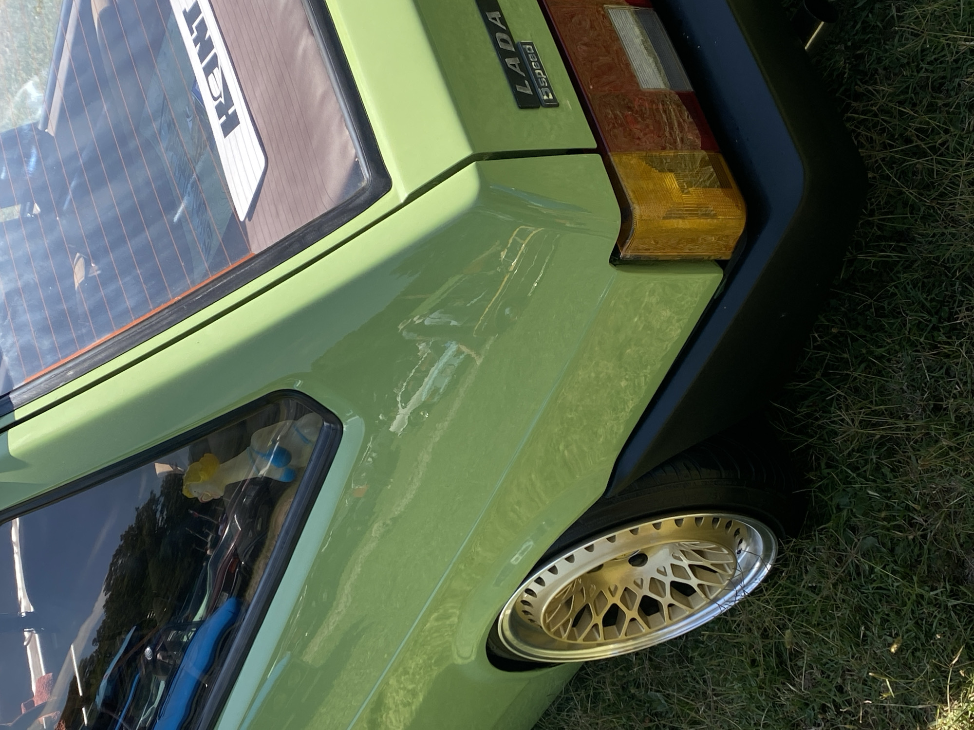 A close-up, eye-level shot captures the rear quarter panel of a bright green, older model Lada car parked on a grassy area. The car's paint is clean and reflects the bright sunlight. Visible on the rear hatch is a white sticker with black lettering that reads "HOITT." A small, plastic figurine of a woman in a blue bikini is visible through the rear window. The car has a gold mesh-style wheel with a chrome lip, and the tire appears to be low to the ground. The rear taillight assembly, with its amber and red lenses, is partially visible. The car is situated outdoors, with green grass covering the ground. The background is blurred, suggesting an outdoor event or parking area, possibly in the city of Bezymyannoe, Russia.