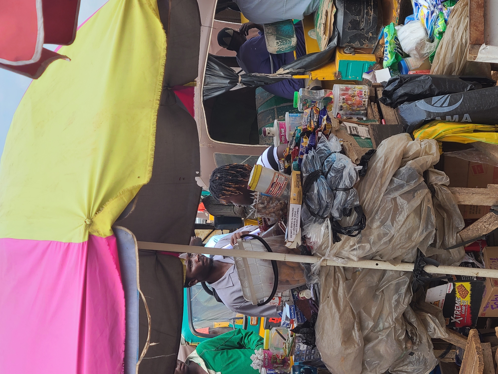 This image captures a vibrant, bustling street vendor scene, likely in Gura Top, Nigeria, given the provided context. The setting appears to be an outdoor market or roadside stall, characterized by colorful textiles and an abundance of goods.
Several individuals are visible, actively engaged in the scene. In the background, a man in a blue shirt and black cap is seated within what looks like a modified vehicle, possibly a tricycle taxi (Keke Napep), serving as part of the mobile shop. Another man, wearing a light-colored shirt and a dark beard, is seen from the chest up, positioned behind a steering wheel-like object, looking directly at the camera. In the foreground, a person with dreadlocks is partially visible, alongside a profusion of merchandise. Further down, another person in a green shirt is present among the stacked items.
The scene is dominated by an eclectic assortment of products, indicative of a general goods vendor. These include numerous packaged snacks, sweets, and drinks, piled high in plastic bags and cartons. Large, brightly colored umbrellas – yellow, pink, and red – provide shade or shelter, common in outdoor trading environments. The overall impression is one of a busy, somewhat makeshift setup, with various items secured by white poles or arranged on shelves made from wood.
Notable details include text visible on various products. "Center fruit" appears on several items, suggesting a brand of confectionery or drink. Other texts include "ORIS," "Aspen," and "XTREME GRAB GO 60 Packets," likely referring to snack brands. A prominent cigarette warning states, "TOBACCO SMOKE CONTAINS OVER 70 SUBSTANCES KNOWN TO CAUSE CANCER." Other discernible words are "Manchester GOLD," "MA," and numbers "22:57," "60544," and "M23 02" on packaging. The bright lighting suggests it is daytime. The cluttered but organized arrangement of goods points to an active trading environment.