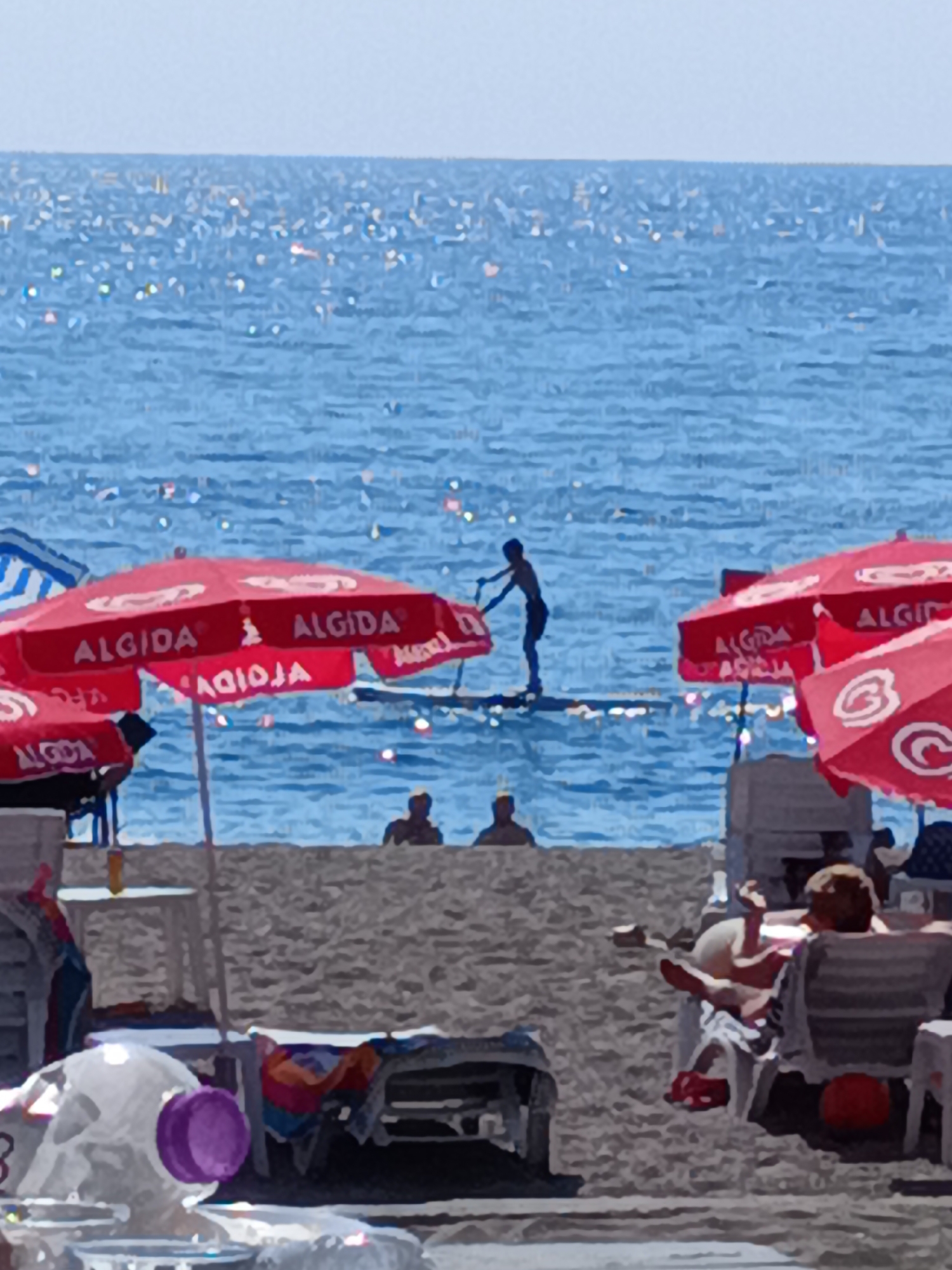 The image captures a sunny day at a beach in Gazipaşa, Turkey. The sea, shimmering under the sunlight, occupies the background. In the middle ground, a person is seen paddleboarding on the water. Red beach umbrellas with the "ALGIDA" logo are prominently displayed on the sandy shore, indicating a commercial establishment. Several beach chairs are scattered on the sand, with some occupied by sunbathers. The foreground shows more beach furniture and some translucent objects, possibly bottles or containers. The overall scene depicts a typical summer day at a seaside resort.