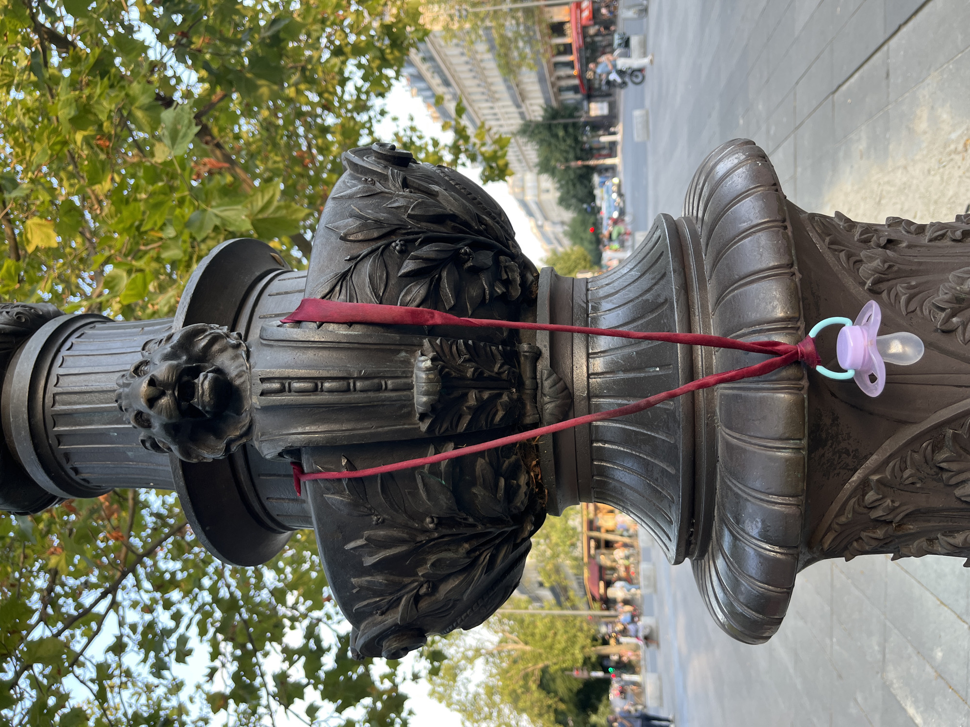 An outdoor, close-up, eye-level, vertical shot shows a decorative lamppost in Paris, France. The lamppost is made of dark metal and features ornate carvings, including a lion's head. A maroon ribbon is tied to the lamppost and dangles down, with a light purple pacifier attached to its end by a light blue ring. The background is slightly blurred, showing a street scene with trees, buildings, and people walking. The sky is visible through the leaves of the trees. The lighting suggests it is daytime, possibly late afternoon.