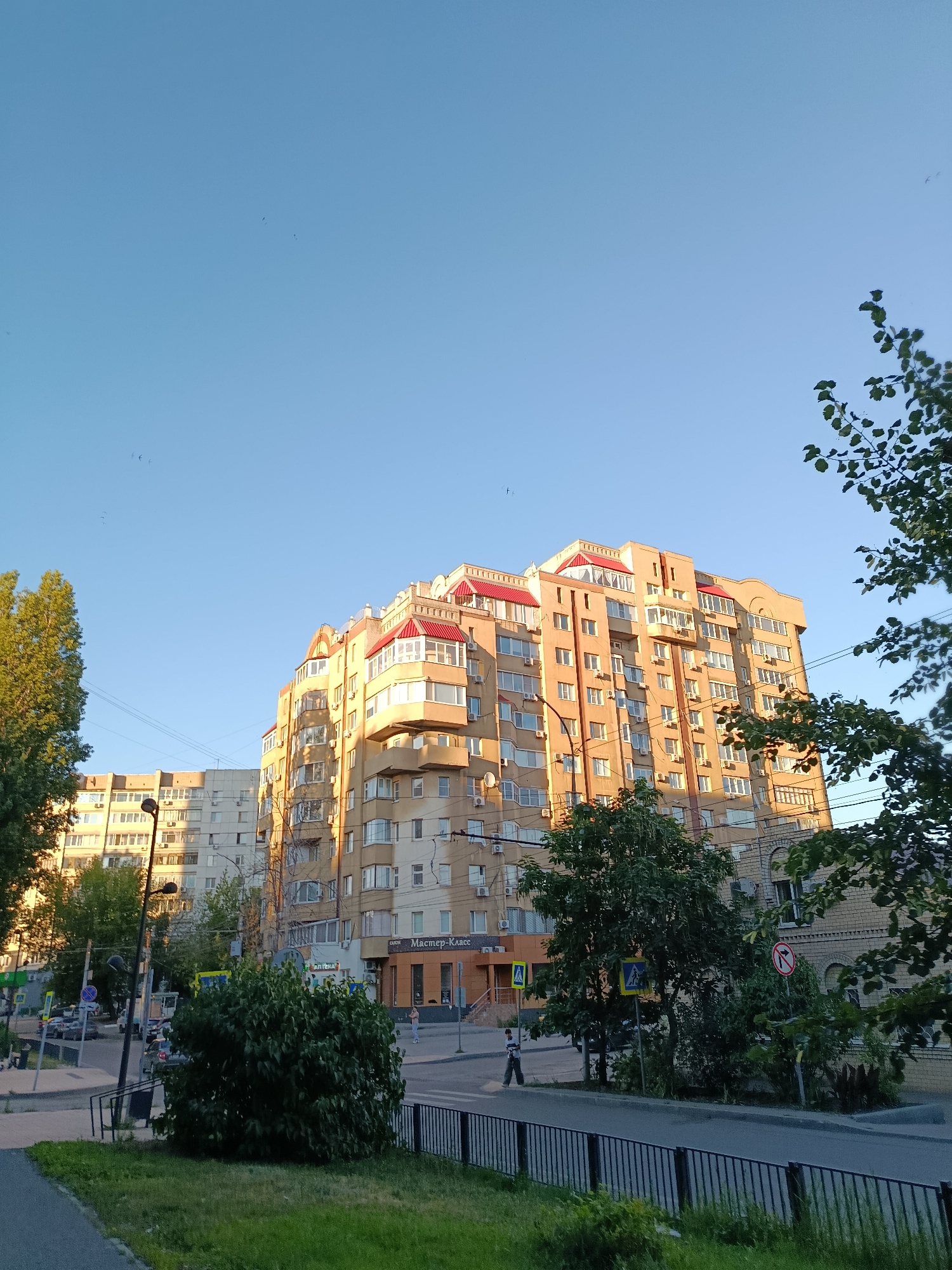 The image depicts a street scene in Saratov, Russia, under a clear blue sky with a warm, late afternoon sun casting long shadows. In the foreground, a black metal fence runs along a strip of green grass and a paved pathway. Beyond the fence, a street with a few parked cars and trees lines the sidewalk. The middle ground is dominated by several apartment buildings. The most prominent is a tall, light-orange building with a red roofline and numerous balconies. To its left, a lighter-colored apartment building stands. In the background, more buildings and trees are visible, along with streetlights and traffic signs. A few birds are visible in the sky. One person is walking across the street in the middle distance. The overall impression is of a quiet urban environment bathed in the golden light of the setting sun.