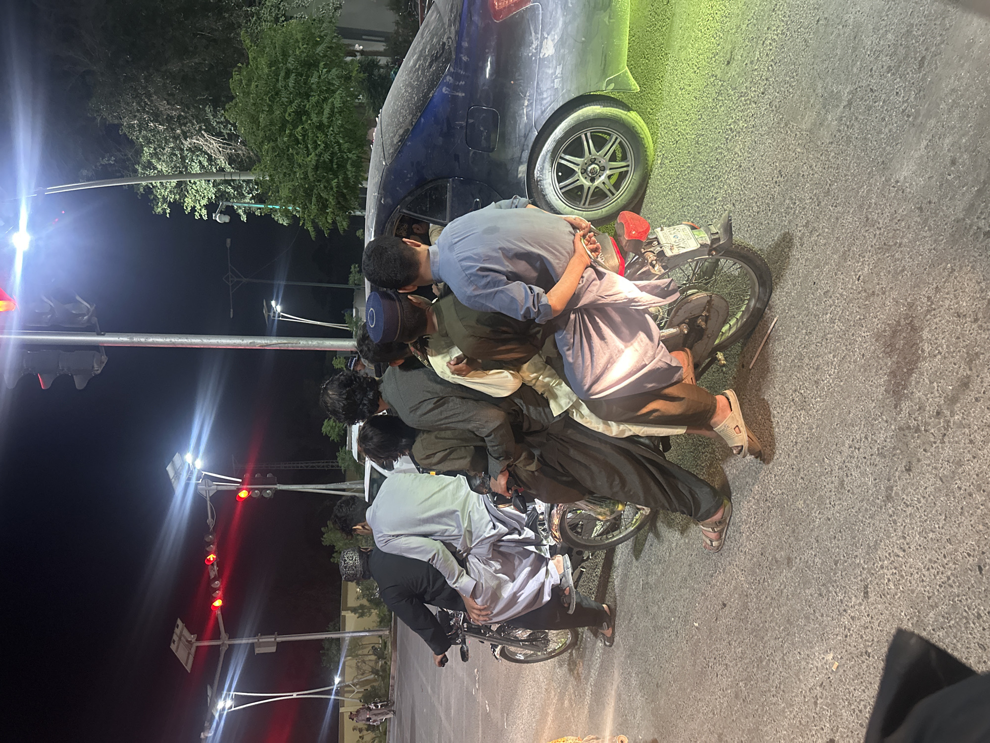 The image captures a nighttime scene on what appears to be a street or roadside. A group of approximately five to six young men, dressed in traditional South Asian attire (shalwar kameez), are gathered around two motorcycles. One man, wearing a light blue shalwar kameez, is leaning over the handlebars of a motorcycle. Another man in a blue cap with a white "O" is positioned behind him, also close to a motorcycle. Their postures suggest they are either conversing, resting, or have momentarily stopped.
To the right of the men, a dark-colored car, possibly a sedan, is parked partially on what looks like a grassy verge and the asphalt road. A distinct green light illuminates the rear quarter of the car and the nearby foliage.
The background features dark skies, indicating it is nighttime. Bright streetlights and traffic lights, prominently displaying red signals, illuminate the scene, creating significant lens flare. Several trees with dense green foliage are visible in the distance and near the car. A surveillance camera is mounted on a pole near some trees. The ground is paved with asphalt. One of the motorcycle license plates appears to have "PAK" and numbers like "46-0937," suggesting a Pakistani setting.