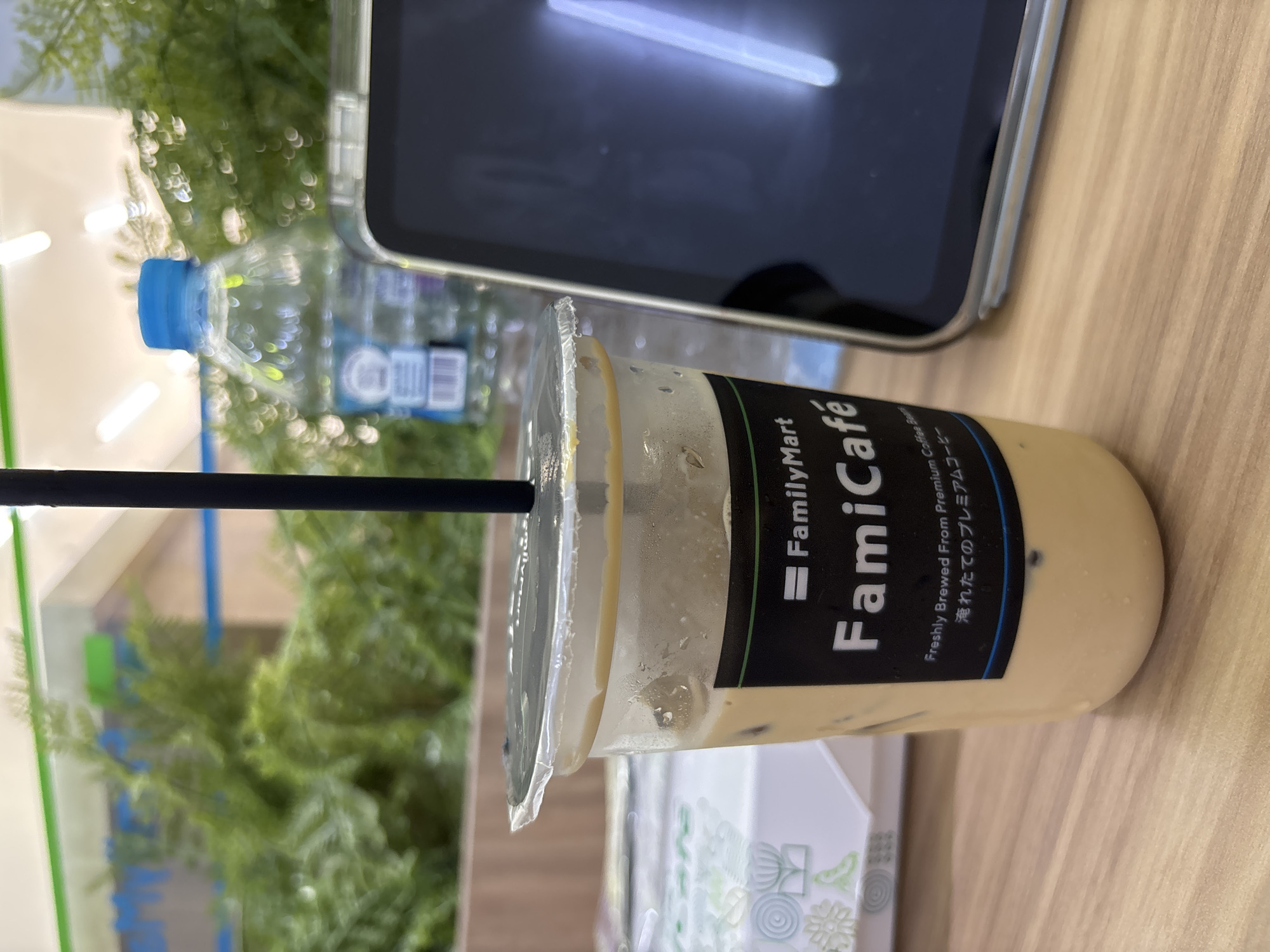 The image, originally rotated ninety degrees to the left, depicts a close-up of a table surface with several items, indicating a moment of rest or work, likely in West Jakarta, Indonesia.
The central subject is a clear plastic cup of iced coffee, positioned prominently. It contains a light brown, milky coffee beverage with ice cubes and a black straw inserted through the clear plastic lid. The cup features a black label with white and green text. Visible text on the label reads: "FamilyMart", "FamiCafé", "Freshly Brewed From Premium Coffee Bea", and in Japanese, "淹れたてのプレミアムコーヒー".
To the left of the coffee cup, a clear plastic water bottle with a blue cap is partially visible. Further back, a dark-screened smartphone or tablet encased in a clear or metallic-rimmed cover rests on the table, reflecting overhead light. The items are placed on a light-colored wooden table surface.
In the background, out-of-focus green foliage suggests either indoor plants or a view through a window to outdoor greenery. A white box or container with faint green and blue patterned designs is partially visible in the bottom right corner of the image. The overall scene suggests a casual setting, possibly a convenience store cafe, a break area, or an outdoor seating area. No people are visible.