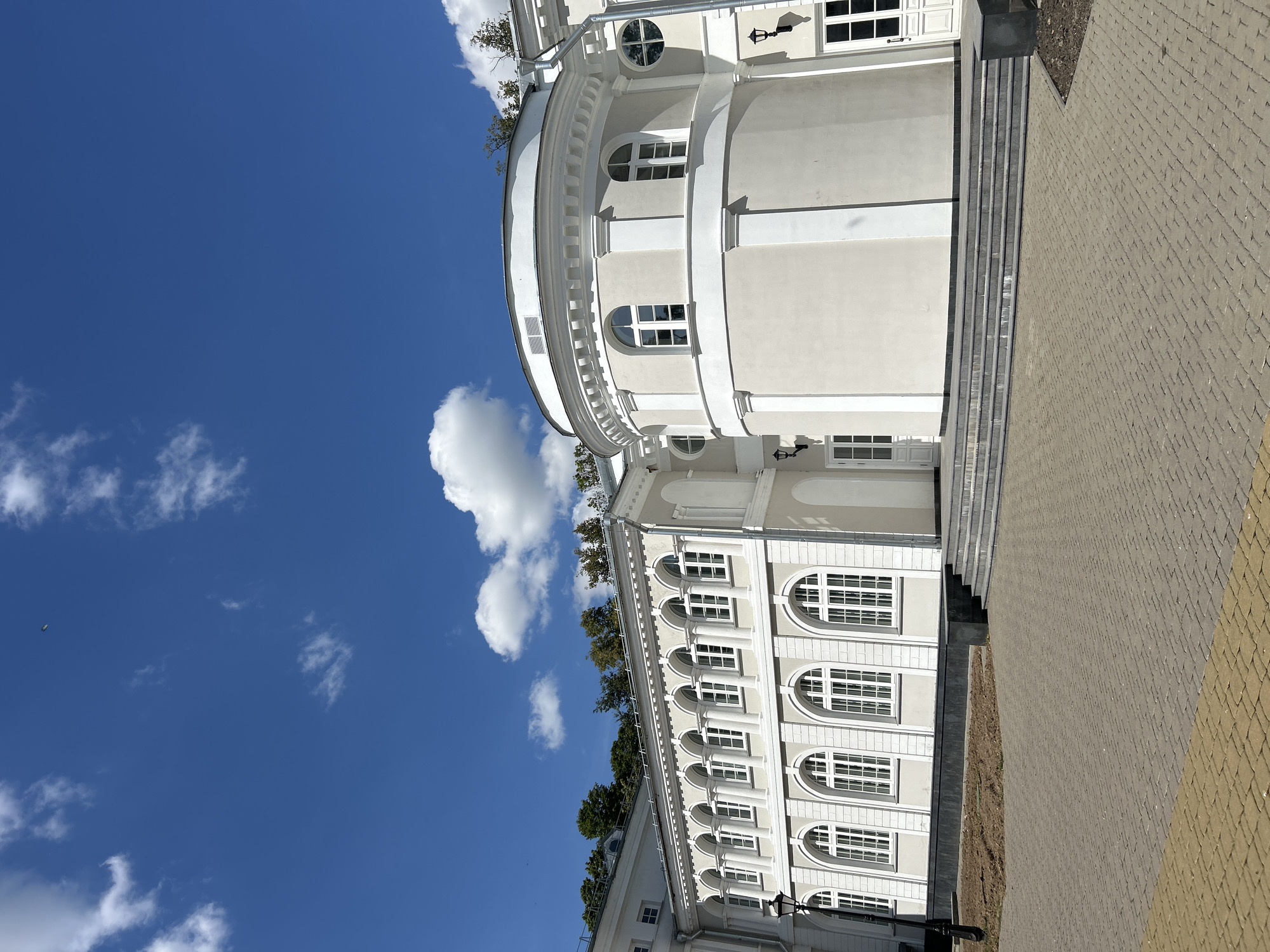 The image shows a light-colored, neoclassical building under a bright blue sky with scattered white clouds. The building features a long, colonnaded section with arched windows and a prominent circular tower-like structure attached to its side. The facade is adorned with decorative moldings and pilasters. In front of the building, there is a paved courtyard and a set of stone stairs leading up to an entrance. Trees are visible along the roofline of the building in the background, suggesting a landscaped setting. A streetlamp stands to the left of the building. The scene appears to be a public or institutional building, possibly a museum, library, or administrative center, located in a well-maintained area. There are no people or animals visible in the image.