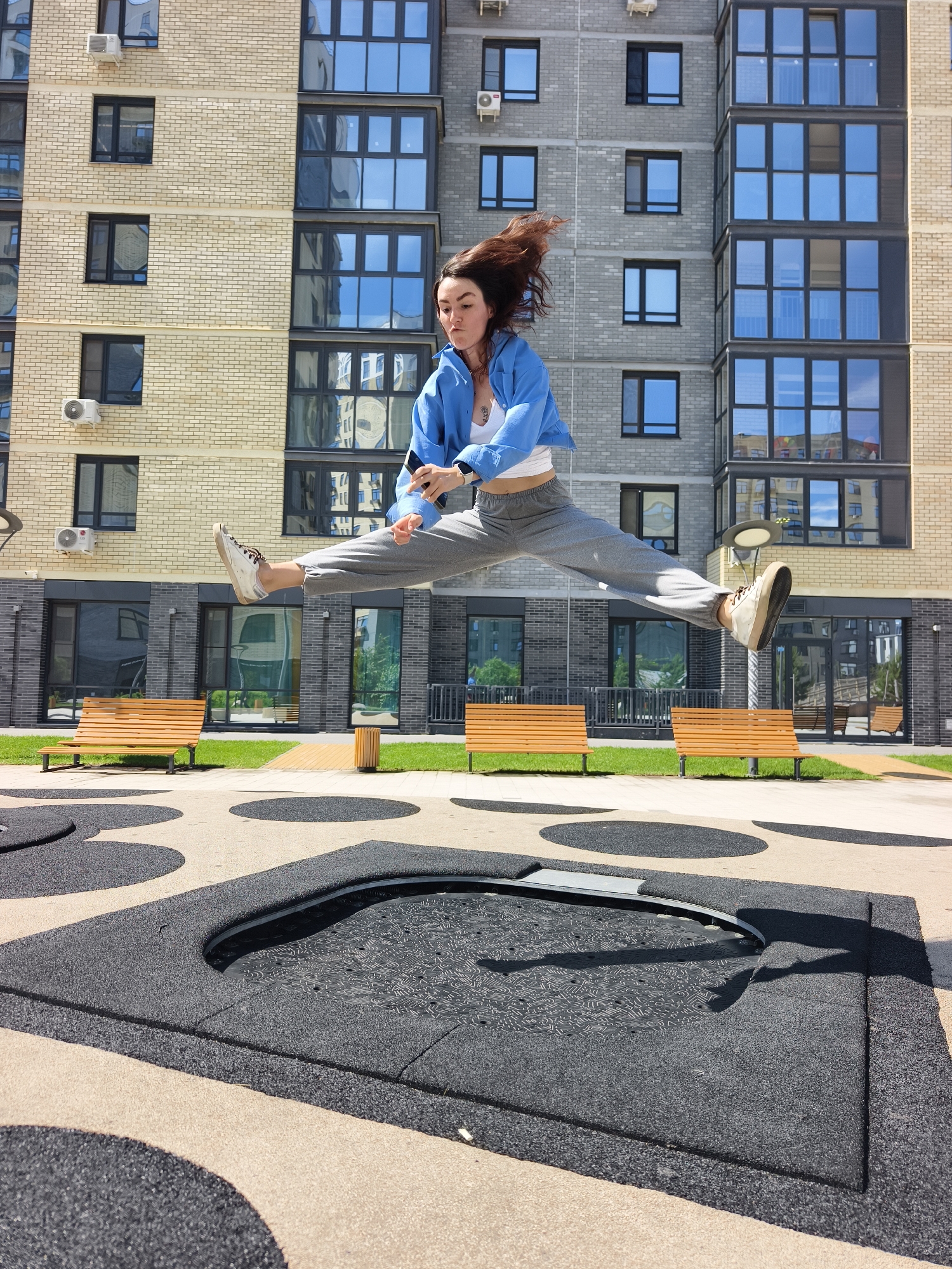 A young woman is captured mid-air performing a dynamic split jump above an in-ground trampoline in an urban courtyard in Tyumen, Russia. She wears a white crop top, grey sweatpants, a light blue unbuttoned jacket, and white sneakers with brown laces. Her brown hair is flying upwards, indicating motion. Her facial expression appears focused.
The trampoline, designed for safety with a textured black surface and a rubberized perimeter, is recessed into the paved ground. Several other black circular patterns are visible on the beige pavement, suggesting an integrated play or recreational area.
In the background, modern residential buildings rise several stories high. The building directly behind the woman features a alternating facade of light-colored brick on the left and darker grey paneling with large, reflective glass windows on the right. Another similar building with a grey facade is visible further back. Several air conditioning units are mounted on the building walls; one unit clearly shows the "LG" logo. The windows reflect the sky and surrounding structures.
Below the buildings, a neatly maintained green lawn is visible, along with several light-colored wooden benches with dark metal frames, indicating a seating area. Strong sunlight casts clear, sharp shadows, suggesting it is a bright, sunny day, likely midday or early afternoon. The overall scene depicts a vibrant outdoor space in a modern residential complex.