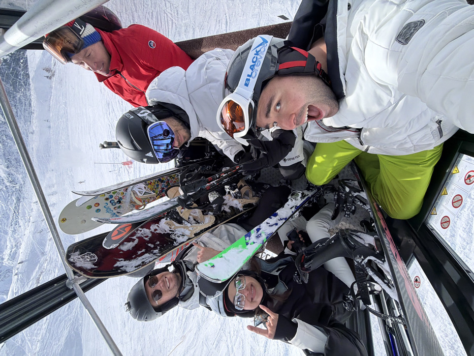 Five individuals are depicted riding inside a ski gondola cabin amidst a snowy, mountainous landscape in Les Allues, France, under bright daylight. The individual in the immediate foreground, wearing a white ski jacket and lime green ski pants, is taking a selfie, mouth agape, and wears a black helmet with orange-lensed goggles. To their left, a female in a black and white ski suit with reflective sunglasses makes a 'rock on' gesture. Another male in a light grey jacket and sunglasses smiles from the upper left. A fourth male, wearing a white jacket and blue-lensed goggles, looks downwards in the mid-right. A fifth male, dressed in a red jacket and a multi-striped beanie featuring an 'LV' logo, looks upwards from the upper right. All individuals are equipped with ski helmets and eyewear. Several snowboards and skis, including a 'Burton' brand snowboard and a pair of green-and-white skis, are stored upright within the cabin. Safety warning labels are visible on the cabin's floor.