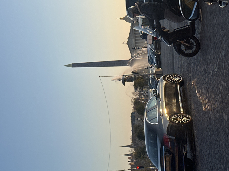 The image depicts a street scene at Place de la Concorde in Paris, France, under conditions suggesting late afternoon or early evening. The sky exhibits a gradient from light blue overhead to a warm yellowish-orange near the horizon.
In the immediate foreground, a person wearing a light-colored helmet and a dark jacket is seated on a black scooter equipped with a clear windshield, positioned on cobblestone paving on the right. To the left, a dark-colored Mercedes-Benz sedan is stationary or moving slowly, with a distinct horizontal reflection of sunlight across its side panel.
In the midground, a prominent fountain is active, discharging multiple streams of water. The Luxor Obelisk, a tall, narrow structure with a distinctive gold-capped pyramidion, stands vertically in the background, offset to the right. To the left of the Obelisk, various Parisian buildings with pointed spires and domes are discernible. A red traffic light is visible on the far left. Farther into the background, a classical building with columns is observed behind the fountain. Multiple other vehicles are visible in the background, indicating active city traffic.