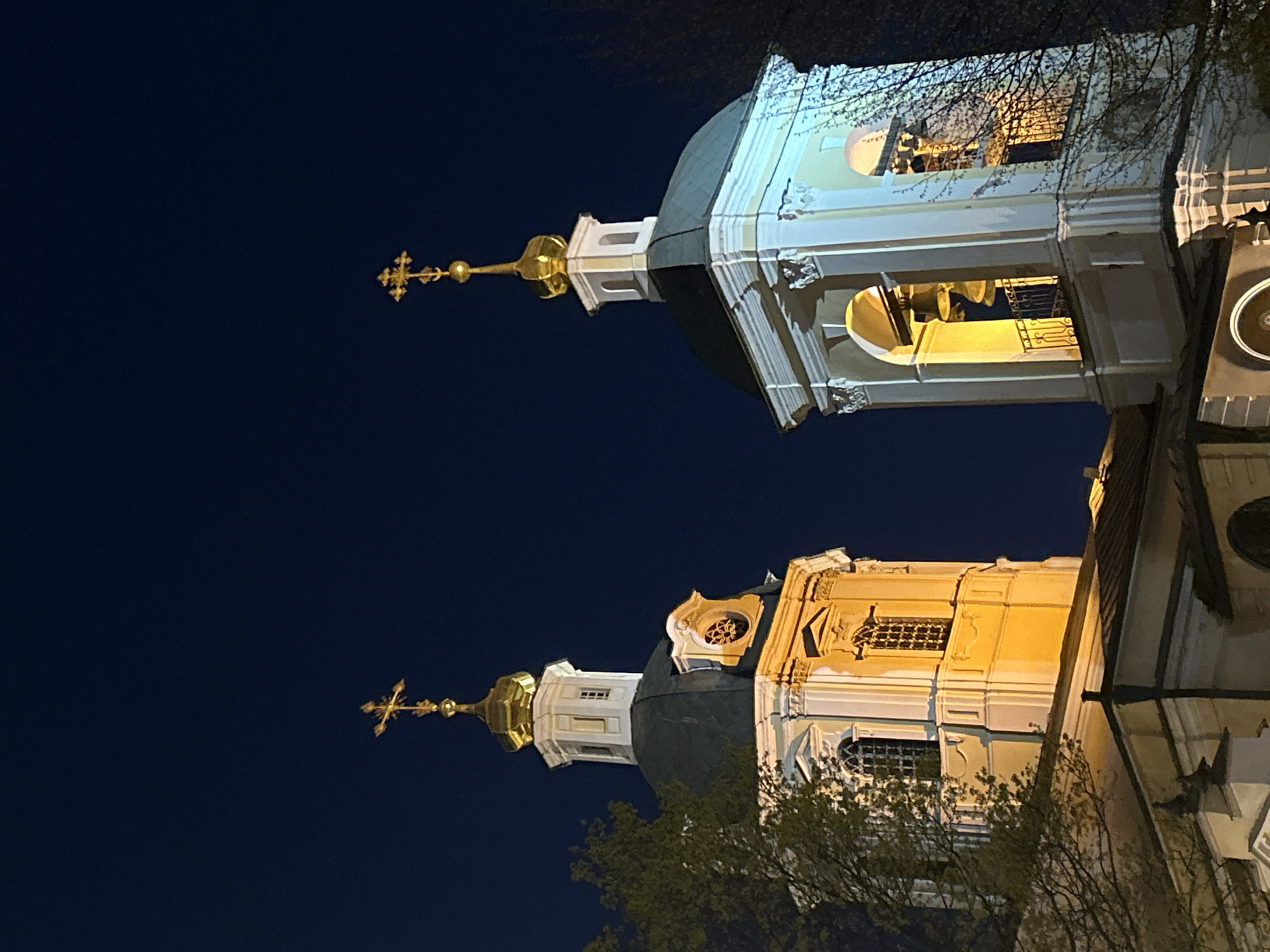 This vertical media file, appearing to be rotated 90 degrees clockwise, captures two brightly illuminated Orthodox church towers or cupolas against a deep dark blue night sky in Moscow, Russia.
In the immediate foreground, on the left side of the image when viewed upright, stands a prominent bell tower or dome. It is crowned with a gleaming golden cross atop a slender spire. Below the cross, a dark, rounded cupola sits on an ornate, multi-tiered structure rendered in white and light blue. This section features several arched openings, inside which large bells are clearly visible, bathed in a cool, almost blue-white artificial light. Bare tree branches are subtly visible, partially framing this structure.
Further back and to the right, a second church tower is visible. This one also features a golden cross and spire above a dark dome. Its architectural details, including decorative carvings, cornices, and arched windows with grates, are illuminated with warmer, golden-yellow artificial light, giving it a distinct appearance from the first tower. More tree branches, some with hints of budding leaves, partially obscure the base of this structure.
The sky is a uniform, deep dark blue, signifying night. The artificial lighting dramatically highlights the architectural details and the golden elements, creating a stark contrast with the surrounding darkness. There are no people visible, nor any discernible activities taking place. No visible text is present in the image. The scene evokes a sense of serene grandeur, showcasing the elaborate and distinctive features of Russian Orthodox architecture under the night sky.