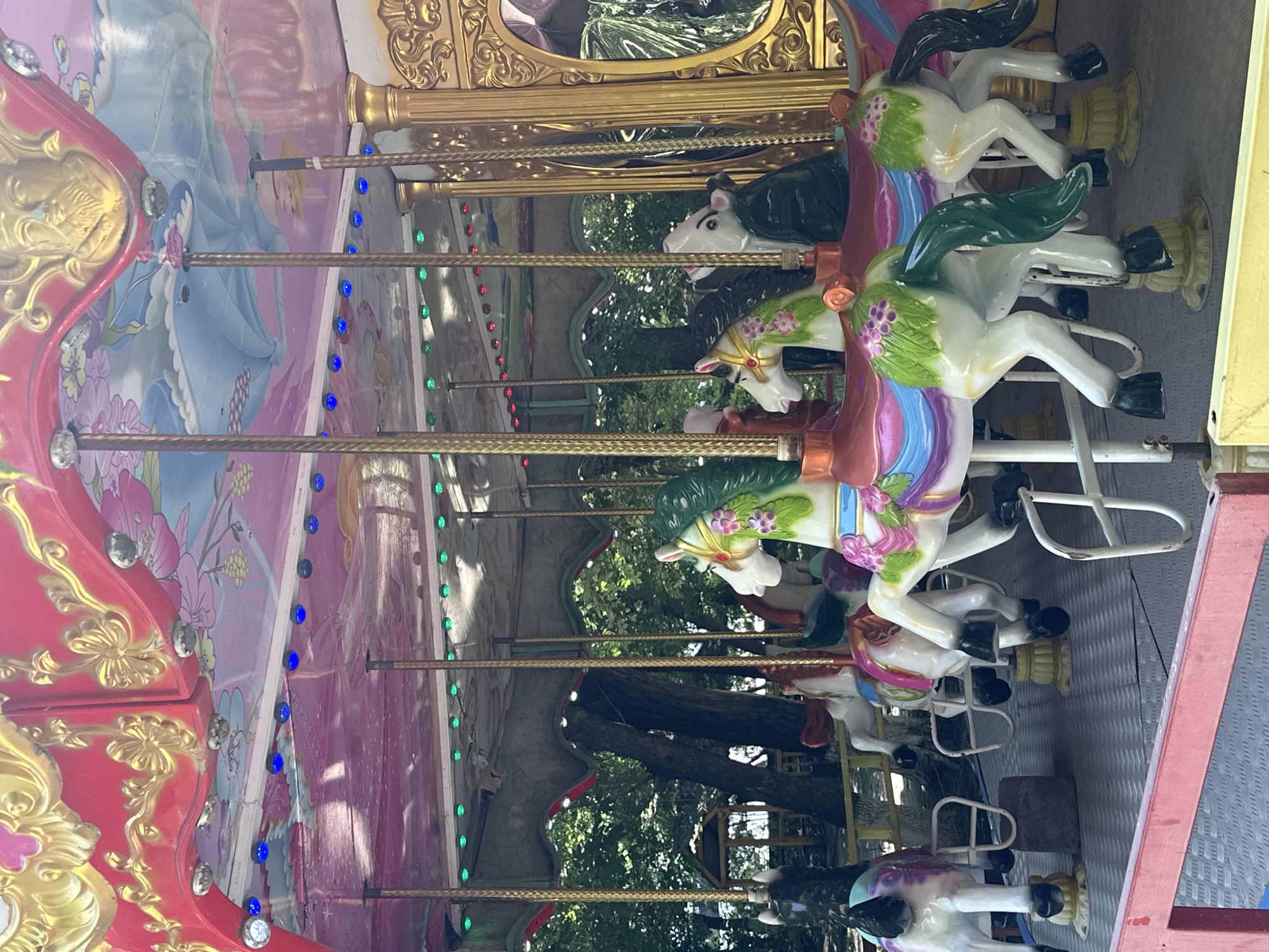 The image shows a carousel with multiple horses. The carousel has a decorative, colorful canopy with intricate designs and lighting. Several horses are visible, each uniquely decorated with vibrant colors and floral patterns. The horses appear to be made of plastic or fiberglass and are mounted on poles. The carousel is situated outdoors, with trees and foliage visible in the background, suggesting a park or amusement area. The ground surface of the carousel platform is a textured gray material, bordered by a red railing in the foreground. There are no people visible in the image. The overall scene depicts a recreational amusement ride. The location in Gyumri, Armenia cannot be determined from the image itself.