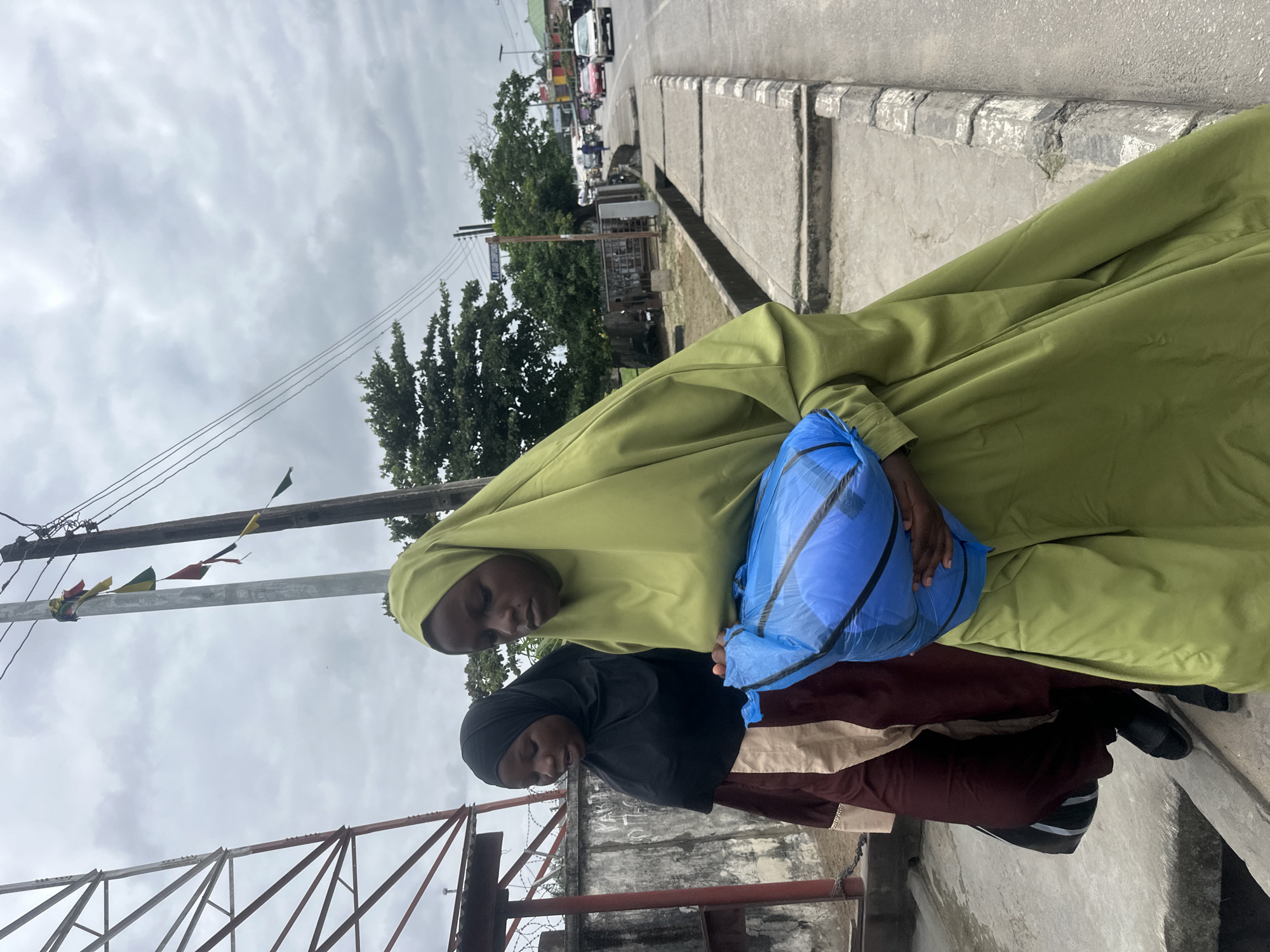 Two young women walk along a paved sidewalk next to a road in Badagry, Nigeria. The woman in the foreground wears a lime green hijab and dress and carries a blue plastic bag filled with what appears to be fabric. She is looking down at the bag. The woman behind her wears a black hijab and a maroon and cream colored dress, and she is looking forward. A chain-link fence is visible behind them, and further in the background are trees and buildings. A utility pole with small, colorful flags hangs above the women. The sky is overcast.
