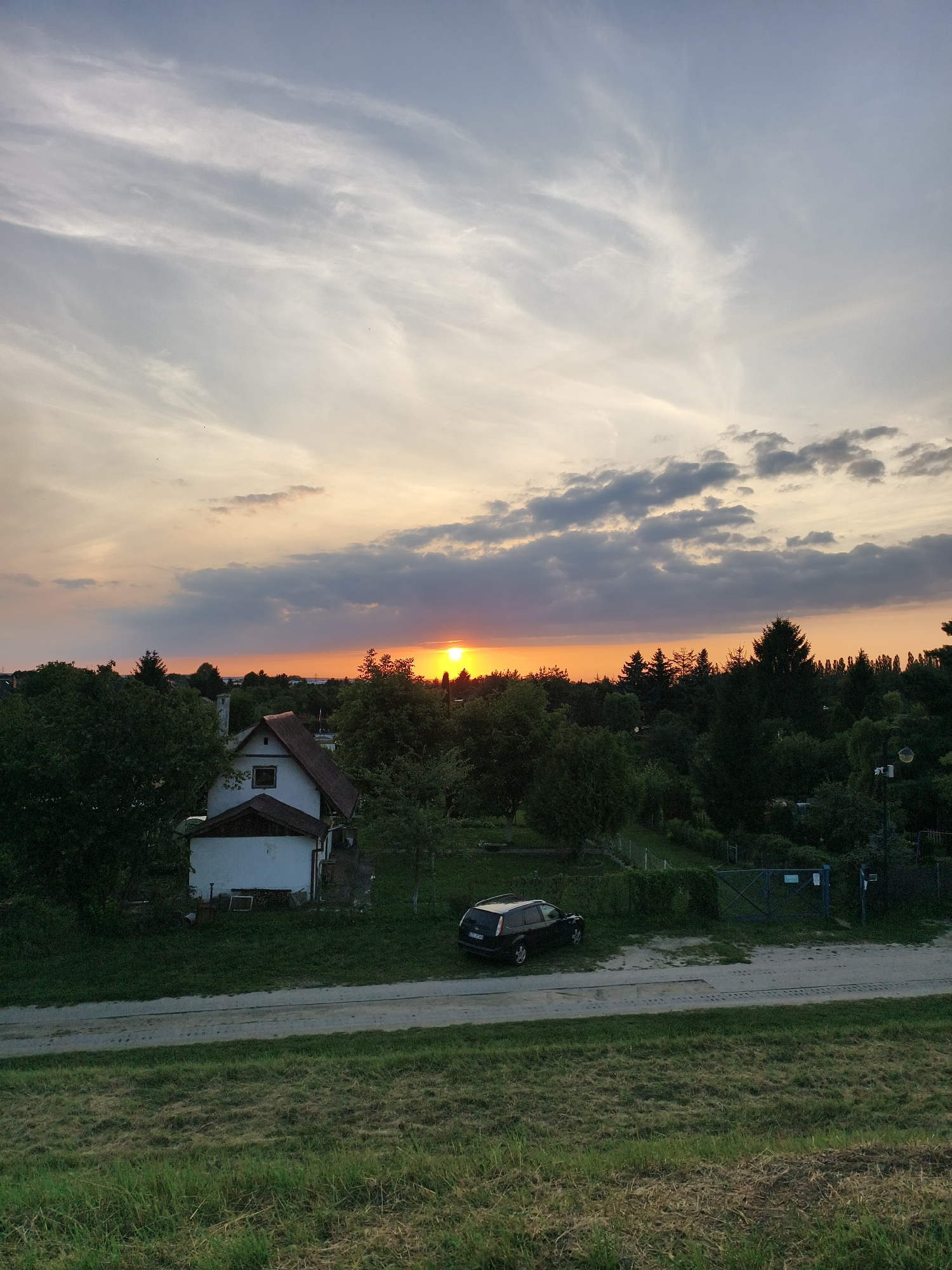 The image captures a sunset over a residential area in Tczew, Poland. The sky is filled with wispy clouds and vibrant orange and yellow hues from the setting sun, which is visible on the horizon amidst a line of trees. In the foreground, a grassy slope leads down to a road. Adjacent to the road is a property featuring a white house with a brown roof, surrounded by lush green trees and vegetation. A dark-colored SUV is parked on the road in front of a fence. The scene depicts a tranquil evening in a suburban setting.