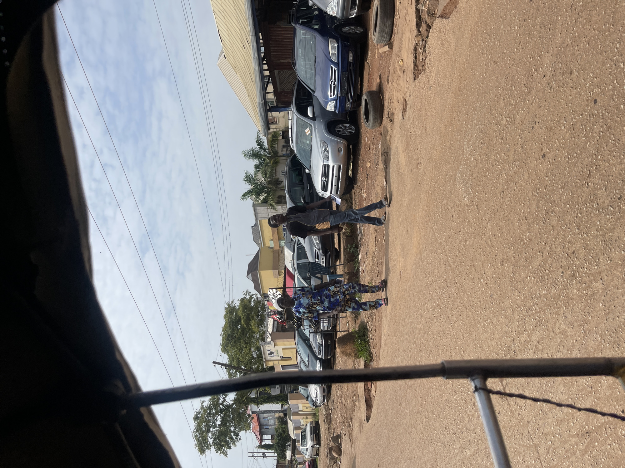 This is a photograph of a street scene in Jos, Nigeria, taken from the perspective of someone inside a tuk-tuk. In the foreground, the metal frame of the tuk-tuk is visible. The street itself is unpaved and dusty, with some rubble and discarded tires scattered around.
Further down the street, a number of cars are parked. These appear to be for sale, with one silver SUV and a blue minivan prominently displayed. Two men are walking on the street. One man, on the left, is wearing a vibrant blue and white patterned traditional Nigerian outfit and a white cap. He is walking away from the camera. The other man, on the right, is wearing a dark t-shirt and jeans and is walking towards the camera. He is holding a small white object in his hand, possibly a piece of paper.
In the background, there are several buildings, some with corrugated metal roofs. Trees are visible, including a large one on the left side of the frame. Power lines crisscross the sky, which is partly cloudy. The overall impression is of a busy but somewhat unkempt urban environment.