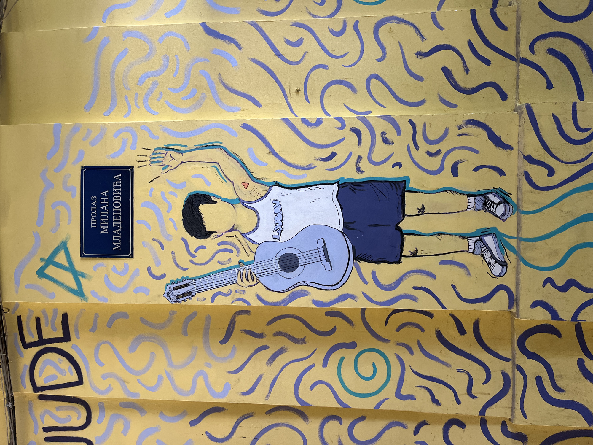 The image depicts a staircase in Novi Sad, Serbia, adorned with street art commemorating Milan Mladenović, the lead singer of the Serbian rock band Ekatarina Velika. The mural features a stylized illustration of Mladenović lying down, holding a guitar, and wearing shorts and a tank top. A small red heart tattoo is visible on his arm. The backdrop is a yellow painted surface with purple and blue wavy lines and spirals. A blue street sign with white Cyrillic text, "ПРОЛАЗ МИЛАНА МЛАДЕНОВИЋА" (Passage of Milan Mladenović), is mounted on the wall. Black graffiti, partially obscured, spells "JUDE" near the bottom of the frame. A blue triangle shape is also painted on the surface near the sign. The artwork covers the steps and wall surfaces of the staircase.