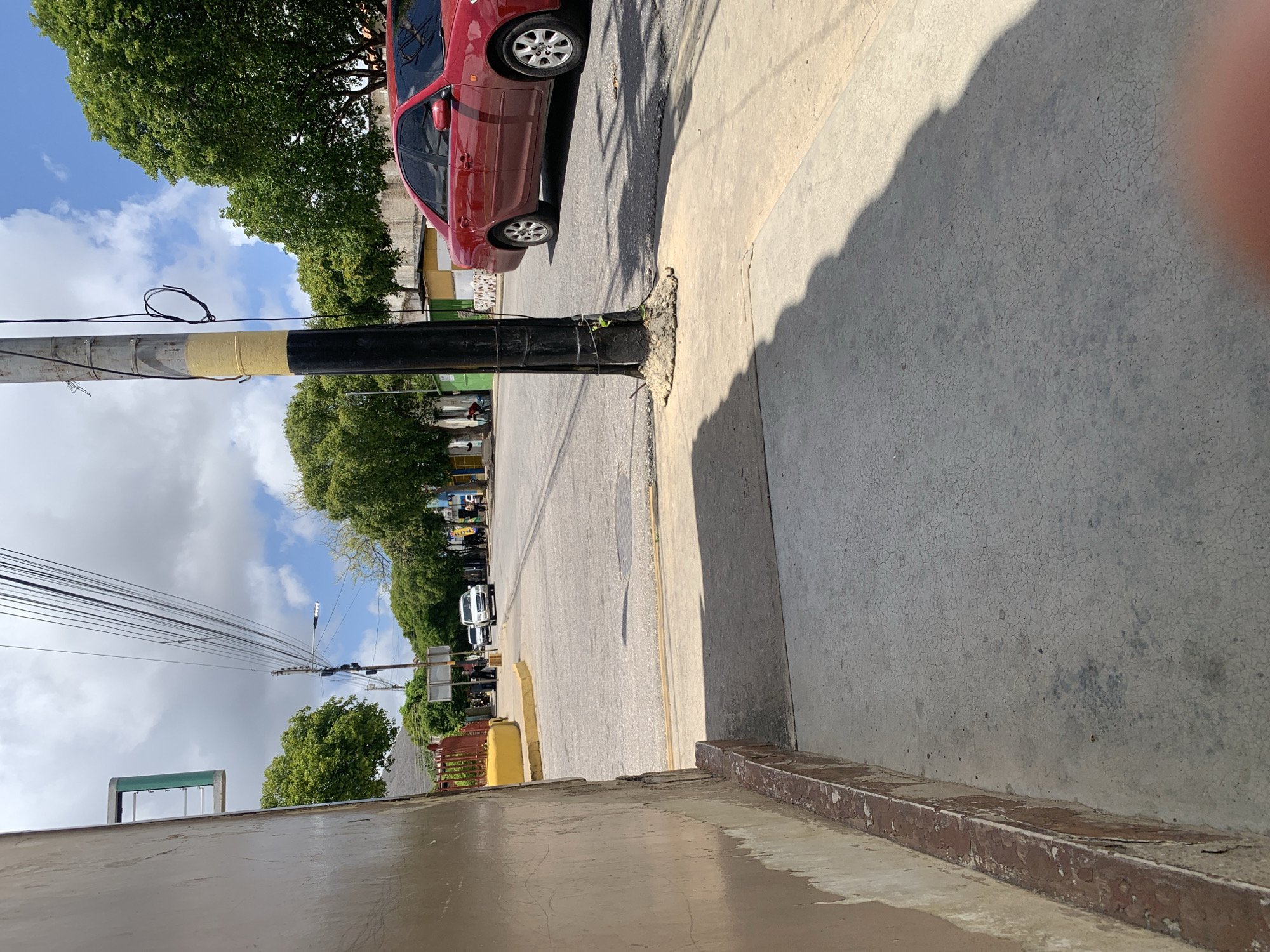 This image depicts a street scene in Santa Ana, Venezuela, on a partially cloudy day. The foreground shows a concrete sidewalk bordering a building with a beige, textured wall and a red-brown trim. To the right, a red sedan is parked or driving on the asphalt street. Beyond the street, a row of green trees lines the road, with buildings and a utility pole visible behind them. Several vehicles are present on the street, including a white SUV further down. The environment suggests a daytime setting with shadows cast by trees and a utility pole.