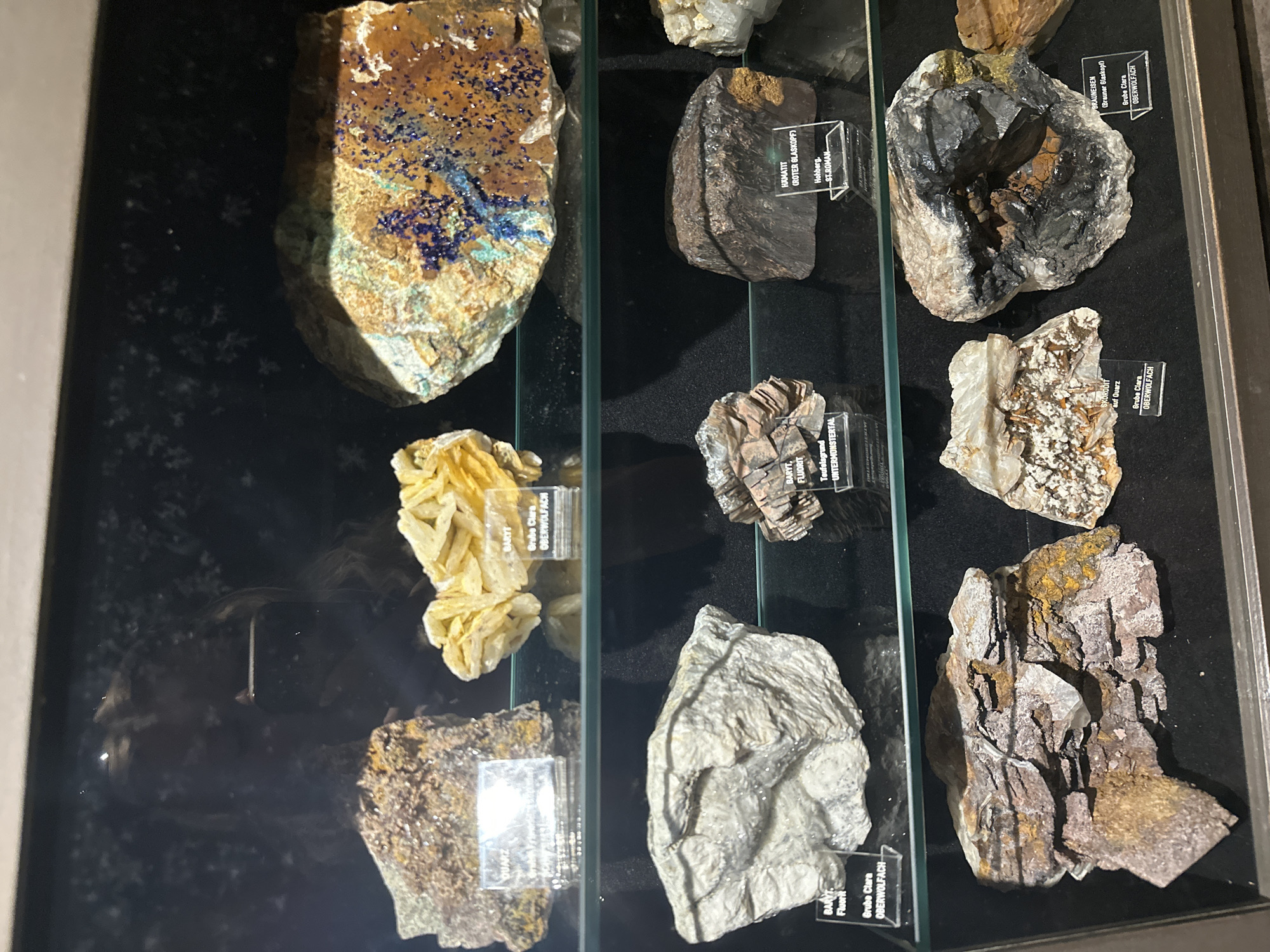 This media file displays a collection of mineral specimens exhibited in a glass-fronted display case. The minerals are arranged in rows within the case, with labels providing information about each specimen. The labels are in German and include terms like "BARYT," "FLUORIT," "HAMATIT," "SKORODIT," and "BRAUNEISEN," along with mine names such as "Grube Clara," "Teufelsgrund," and "Hohberg." The location context provided is Schonach im Schwarzwald, Germany, which suggests these minerals may be from the Black Forest region. The lighting within the display case is artificial, creating reflections on the glass. The background is dark, making the minerals stand out. The overall scene depicts a museum or geological exhibit showcasing a variety of rock and mineral samples. There are no people or active events depicted; the focus is solely on the static display of the mineral collection.