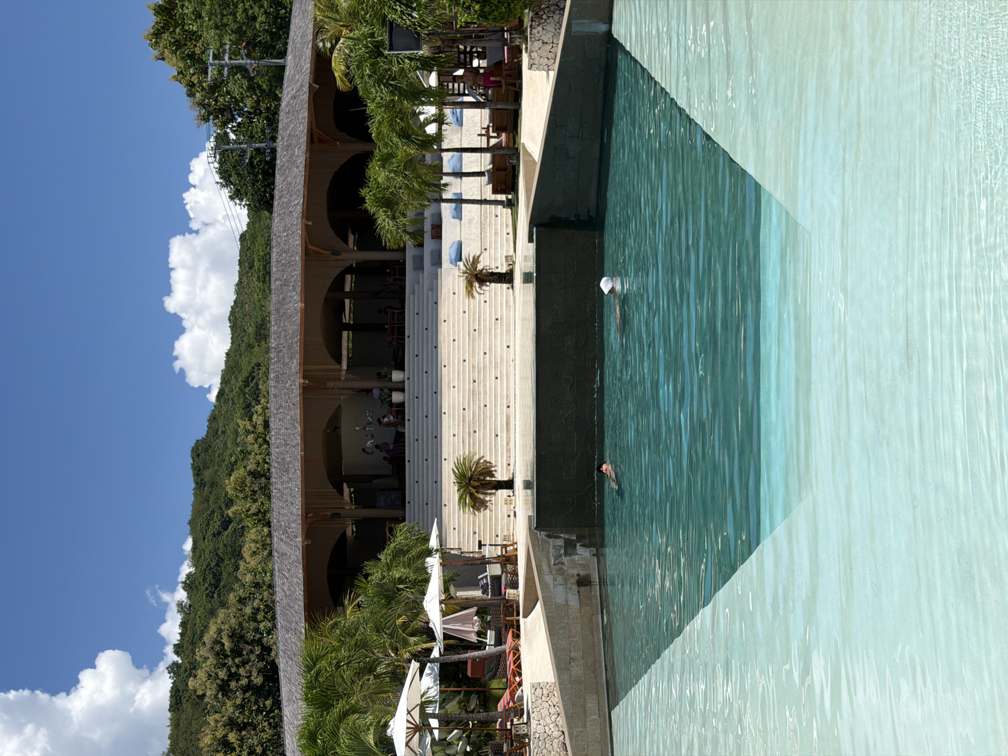 The image depicts a luxurious resort in Batununggal, Indonesia, featuring a multi-tiered swimming pool with clear turquoise water. The pool extends from a concrete platform with decorative palm trees, a staircase, and seating areas shaded by white umbrellas. Adjacent to the pool, a building with arched openings and a dark-colored roof is visible, possibly a restaurant or lounging area. Lush green vegetation covers the hillside in the background, contrasted by a clear blue sky with scattered white clouds. Two individuals are seen swimming in the pool.