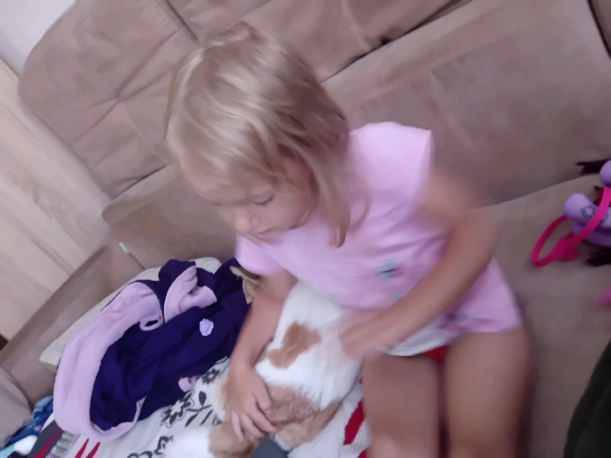 An overhead, slightly blurry, medium shot shows a young Caucasian girl with blonde hair sitting on a beige sofa. The girl is wearing a pink t-shirt and red bottoms. She is holding a stuffed animal with brown and white fur. To her left are piles of folded clothing, including a purple and light pink robe. To her right, on the sofa, is a purple toy with a pink strap. The background is a textured beige sofa. The image appears to be taken indoors, possibly in a residential setting in Chuhuiv, Ukraine, given the provided context, though there are no specific landmarks or indicators to confirm this location within the image itself.