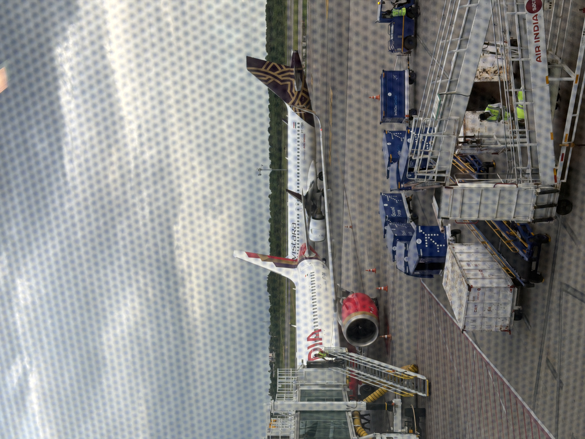 The image, taken from an elevated perspective through a mesh-covered window, depicts an airport scene in Mamidipally, India. A Vistara aircraft, white with red engine nacelles and a maroon and gold tail fin, is parked at a gate, connected to a jet bridge. Several airport service vehicles and cargo containers are visible on the tarmac. Two individuals in high-visibility vests are present, one near the jet bridge stairs and another operating a ground support vehicle. The sky above is overcast with a pattern of hexagonal dots superimposed over the entire image due to the viewing medium.