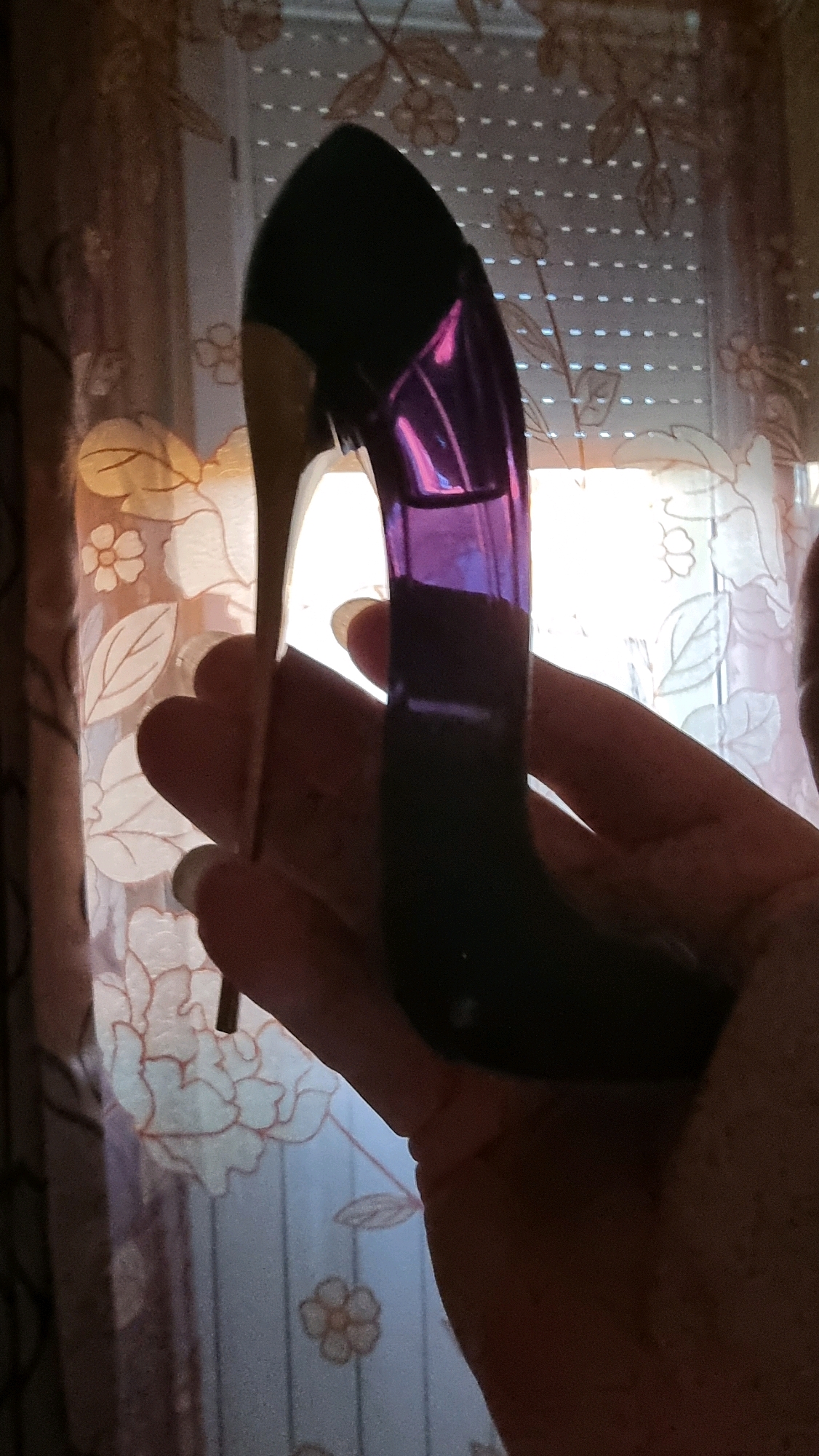 A hand is holding a high-heeled shoe shaped perfume bottle in front of a window with blinds and a floral patterned curtain. The perfume bottle is black and iridescent purple. The background is dimly lit with light coming from the window, revealing a blurred floral pattern on the curtain and vertical blinds. The scene is indoors, suggesting a domestic setting in Sant Joan Les Fonts, Spain.