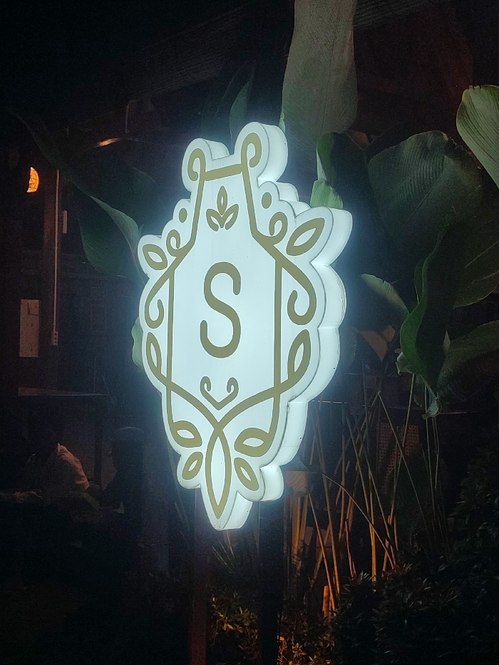 A lit, white sign with a gold 'S' in the center and ornate, gold, floral designs around it is mounted to a pole in front of dense foliage. The sign is brightly lit and appears to be a storefront sign. The sign is located at the coordinates -7.747, 112.737 in the city of Purwosari, Indonesia. The foliage surrounding the sign appears to be a variety of green plants. The scene is dark except for the lit sign.