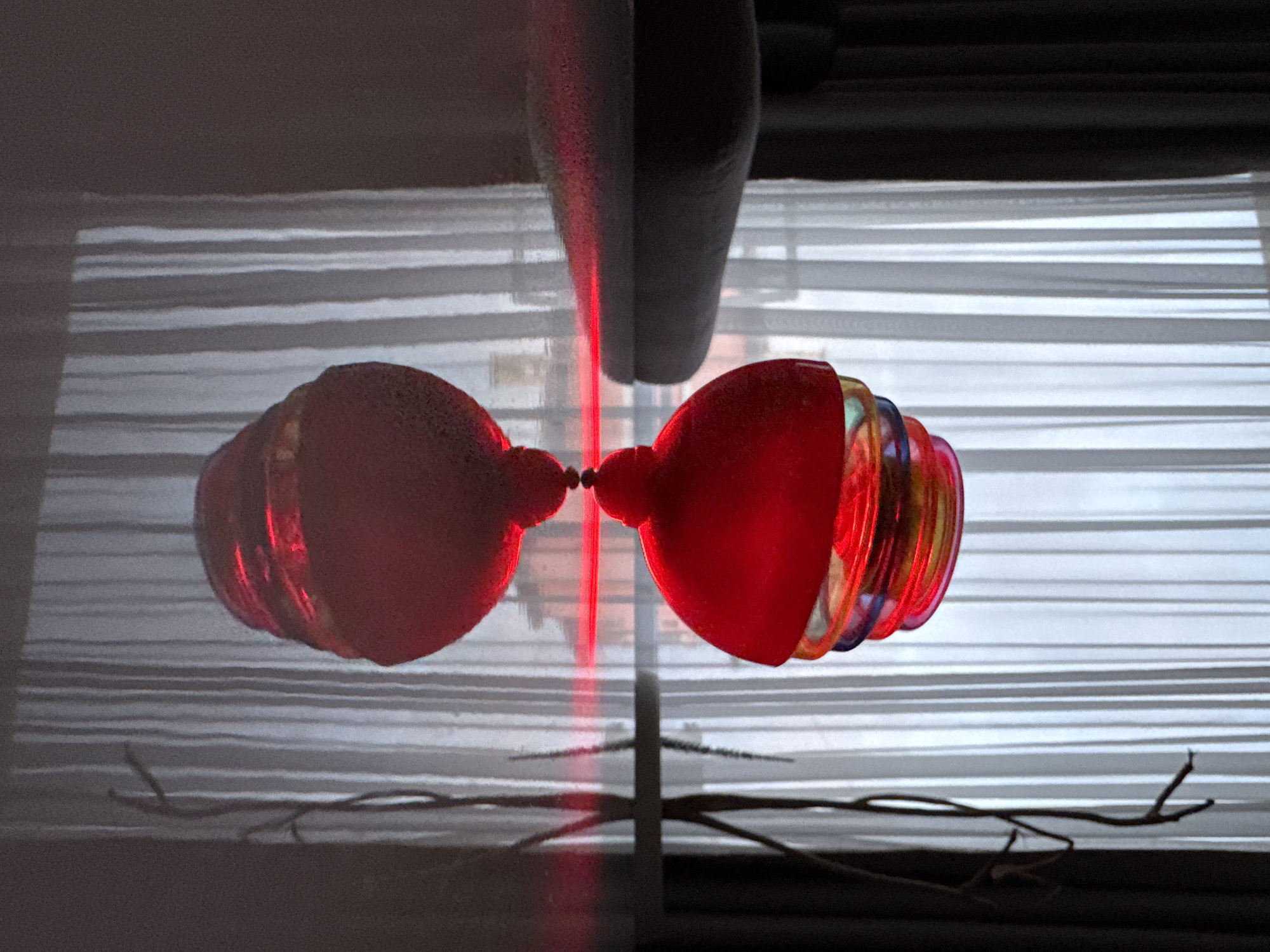 The image displays a highly reflective indoor scene, likely captured in Varna, Bulgaria, characterized by strong light and symmetry. Central to the composition is a vibrant, ruby-red, bulbous object, possibly a decorative ornament or glass bauble, which appears to be composed of stacked rings that reveal hints of blue, yellow, and green where light refracts through them. This object is bisected by a sharp, vertical beam of intense red light, strongly suggesting a laser, which creates a prominent line dividing the reflected image.
The entire scene, including the red object and the red light beam, is mirrored, implying the photograph was taken reflecting off a polished surface, such as a tabletop, or through a pane of glass. In the background, horizontal blinds or window slats are clearly visible, casting striped patterns of light and shadow, suggesting an interior space looking out a window. In the foreground, below the main red object, a dark, gnarled branch or piece of wood rests on a surface, also seen in reflection. Dark, indistinct shapes occupy the upper and lower portions of the image, likely elements of the room or window frame. The dominant red color of the object and light contrasts with the cooler gray and white tones of the blinds and the dark elements. No people or visible text are present. The time of day and weather cannot be determined from the image.