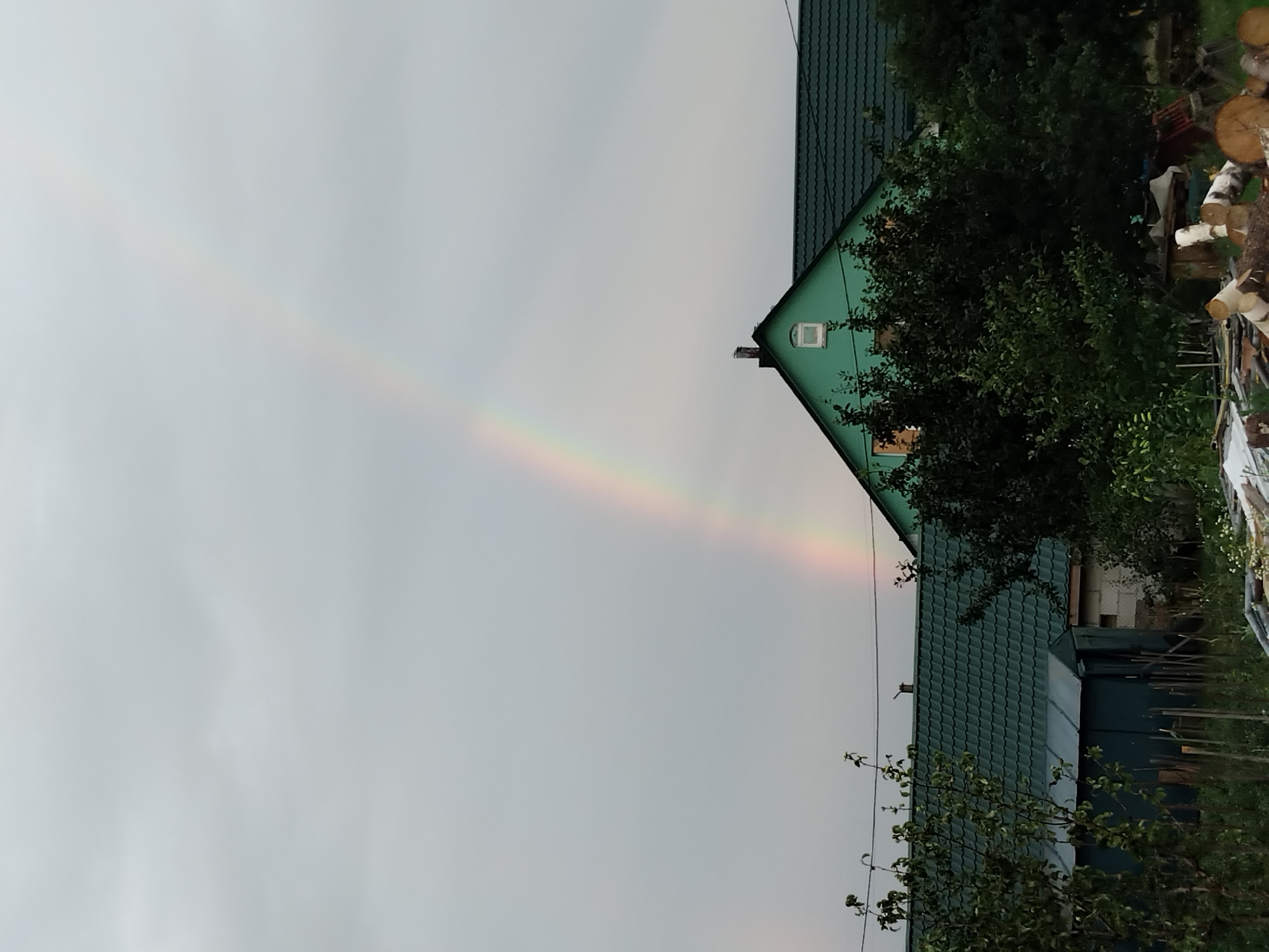 The image captures a rural scene under a cloudy sky with a faint rainbow visible. In the foreground, there are trees, a small shed, and a pile of logs. In the background, a green house with a dark green roof is visible. The house has a chimney and a small, white, arched window. The overall impression is one of a peaceful, natural environment, possibly a backyard or a farm.