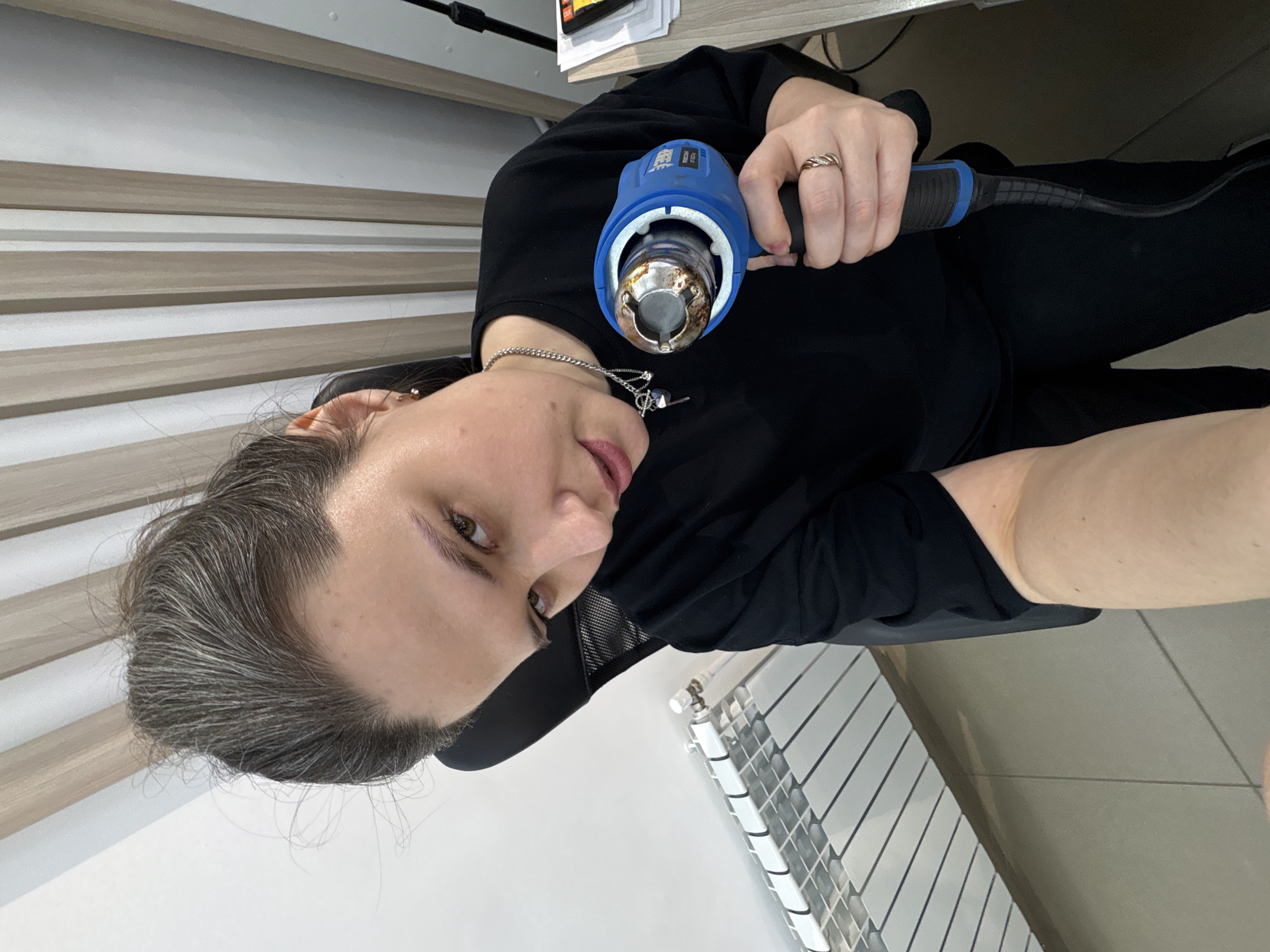 An adult female, with dark hair interspersed with gray strands pulled into an updraft, directly faces the camera from an indoor location in Chaikovsky, Russia. She is seated in a black office chair, wearing a black short-sleeved top and black pants. A silver-colored chain necklace with multiple small charms is visible around her neck, along with a small earring in her left ear. A gold-toned, multi-band ring adorns her right ring finger. Her right hand firmly grips a blue and black "TREP" branded heat gun, its metal nozzle discolored from use, oriented upwards and to the left. Her left forearm is partially visible towards the lower-left frame. The background features a white wall with a white panel radiator on the left, transitioning to vertical alternating light wood-grain and white slatted panels on the right. A light wood-grain desk surface, holding a stack of papers and a black stand, is visible in the lower right corner.