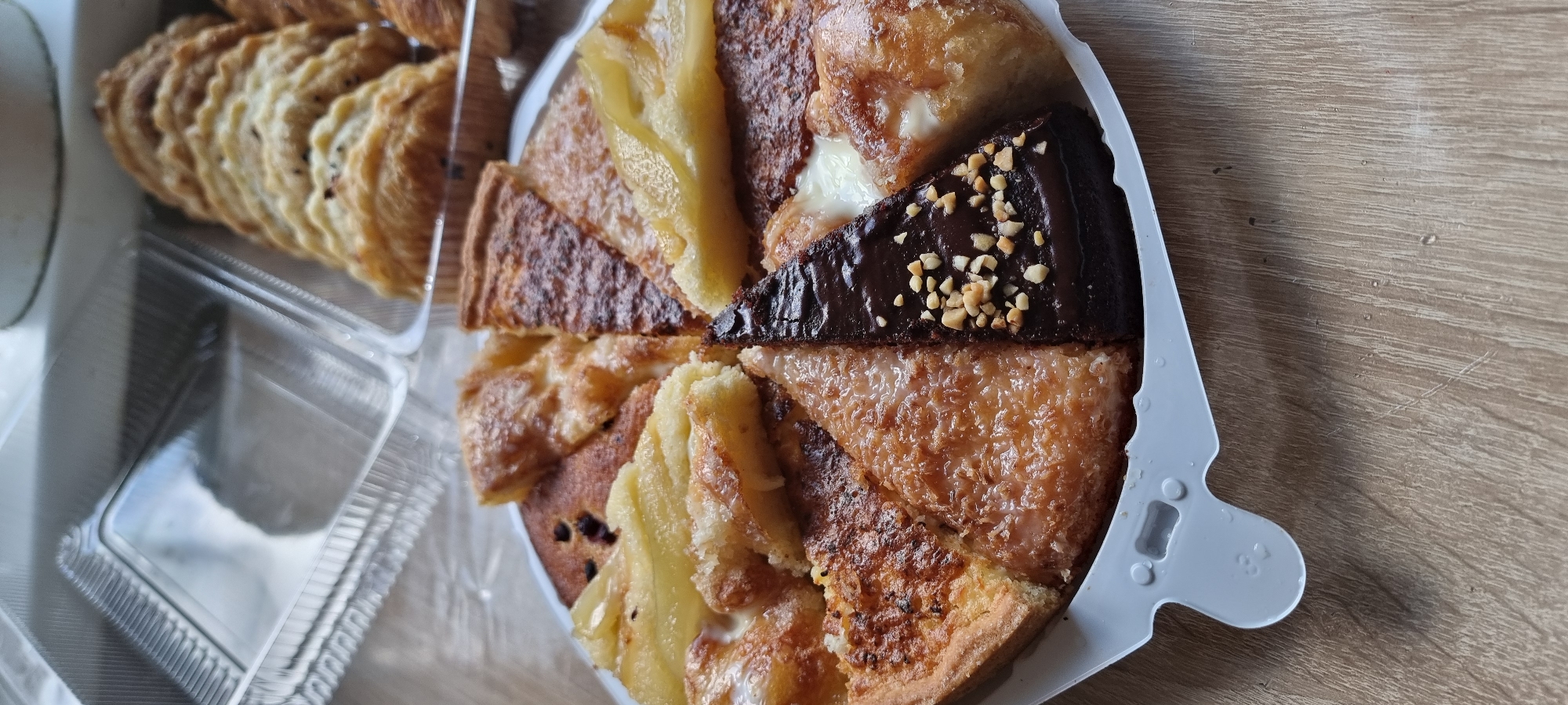 A circular pie, cut into multiple triangular slices, is presented in a white plastic container on a wooden surface. The pie features various toppings and fillings, including what appears to be apple slices, a chocolate glaze with chopped nuts, and a textured topping resembling shredded coconut. Behind the pie, a clear plastic container with a ribbed texture is visible, and further back, a tray of pastries, possibly croissants or turnovers, is partially visible. The wooden surface shows faint grain patterns and a few small water droplets. The scene is lit from above, casting soft shadows. The overall impression is of a selection of baked goods displayed for sale or consumption.