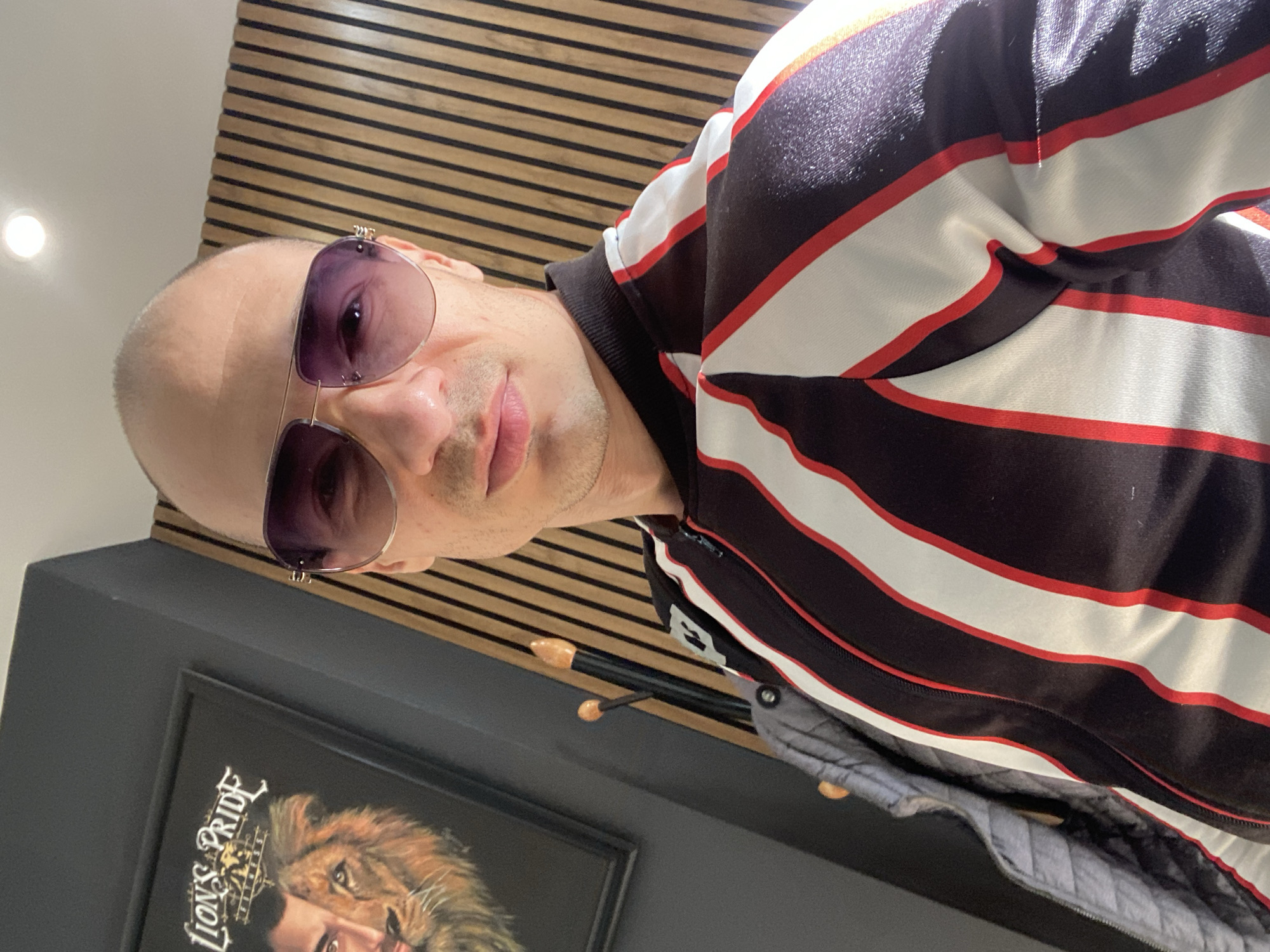 A man with a shaved head is wearing sunglasses and a black, white, and red striped shirt. He is standing in front of a wall with horizontal wooden slats. To the left, there is a framed picture with the words "Lion's Pride" and an image of a lion. A gray quilted jacket hangs on a coat rack below the slats. The location is most likely indoors, in Pleven, Bulgaria, given the prompt's specification. The man is facing the camera and has a neutral expression.