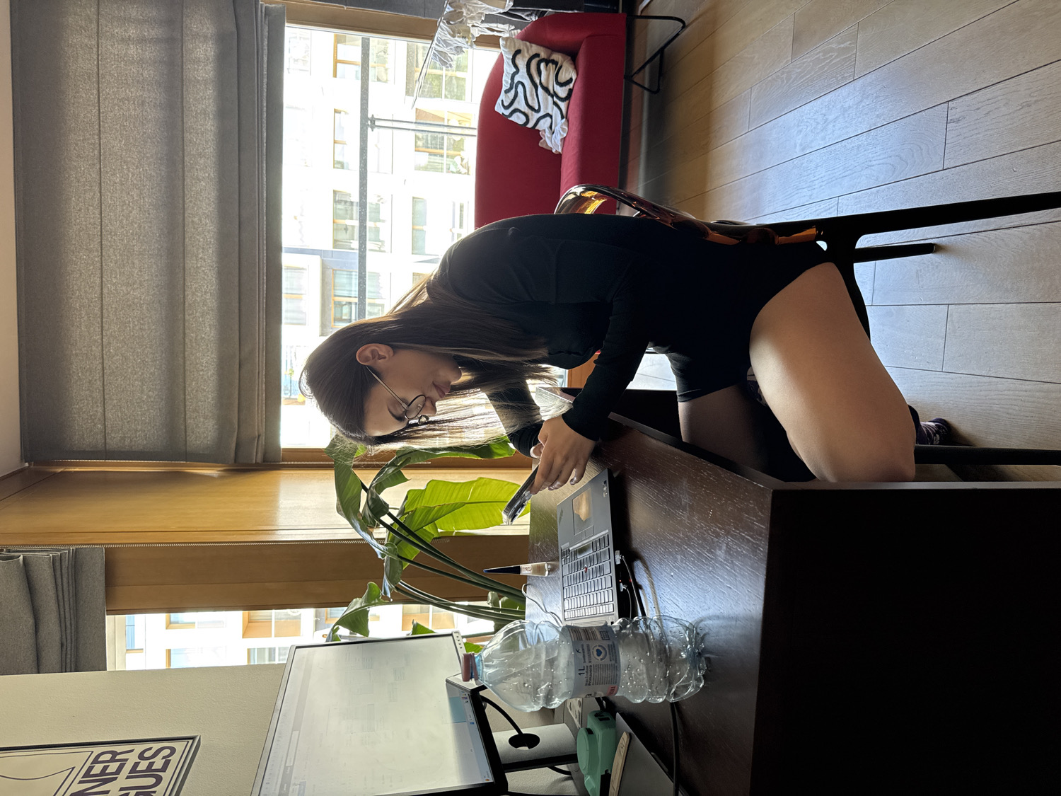 The image depicts an indoor scene, likely a modern apartment or workspace, characterized by ample natural light streaming through a large window. The primary subject is a person, appearing to be a woman, with long dark hair and glasses, dressed in a black long-sleeved top and black shorts or a bodysuit. She is positioned on her knees, leaning forward over a dark wooden desk.
On the desk, a silver laptop, resembling an Apple MacBook, is open and appears to be in use, with her hands near the keyboard. Adjacent to the laptop is a clear plastic water bottle, marked "1L". A large green leafy plant with prominent leaves is positioned near the desk, adding a touch of nature to the setting. In the foreground, partially visible on the left, is another computer monitor and a framed poster with fragmented text reading "...NER GUES".
The room features a light wooden floor and grey vertical blinds covering part of the large window. Through the window, modern apartment buildings are visible under bright daylight conditions, suggesting it is daytime and possibly sunny. In the background, a red armchair with a black and white patterned cushion is discernible, along with part of a black chair or stool with a metallic frame. The overall atmosphere suggests a focused activity, possibly work or study, taking place in a well-lit, contemporary environment in Warsaw, Poland.