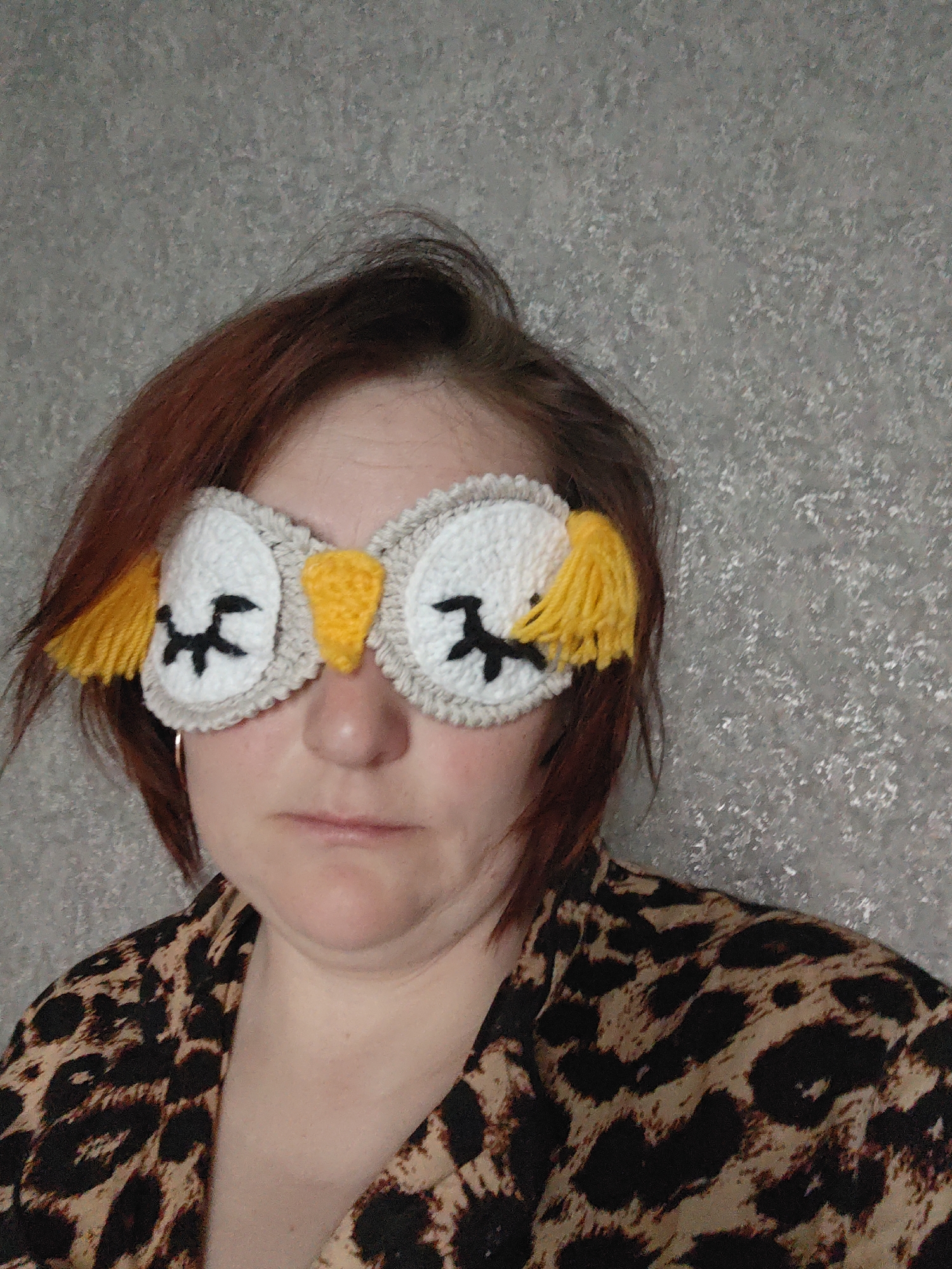 A woman is wearing an owl-shaped sleep mask. The mask is crocheted with white eye patches, black stitched eyes, a yellow beak, and yellow tassels on the sides. She is wearing a leopard-print top. Her hair is reddish-brown and somewhat disheveled. The background is a grey wall with a textured pattern. There is no evidence to suggest the location is Markovskii, Russia.