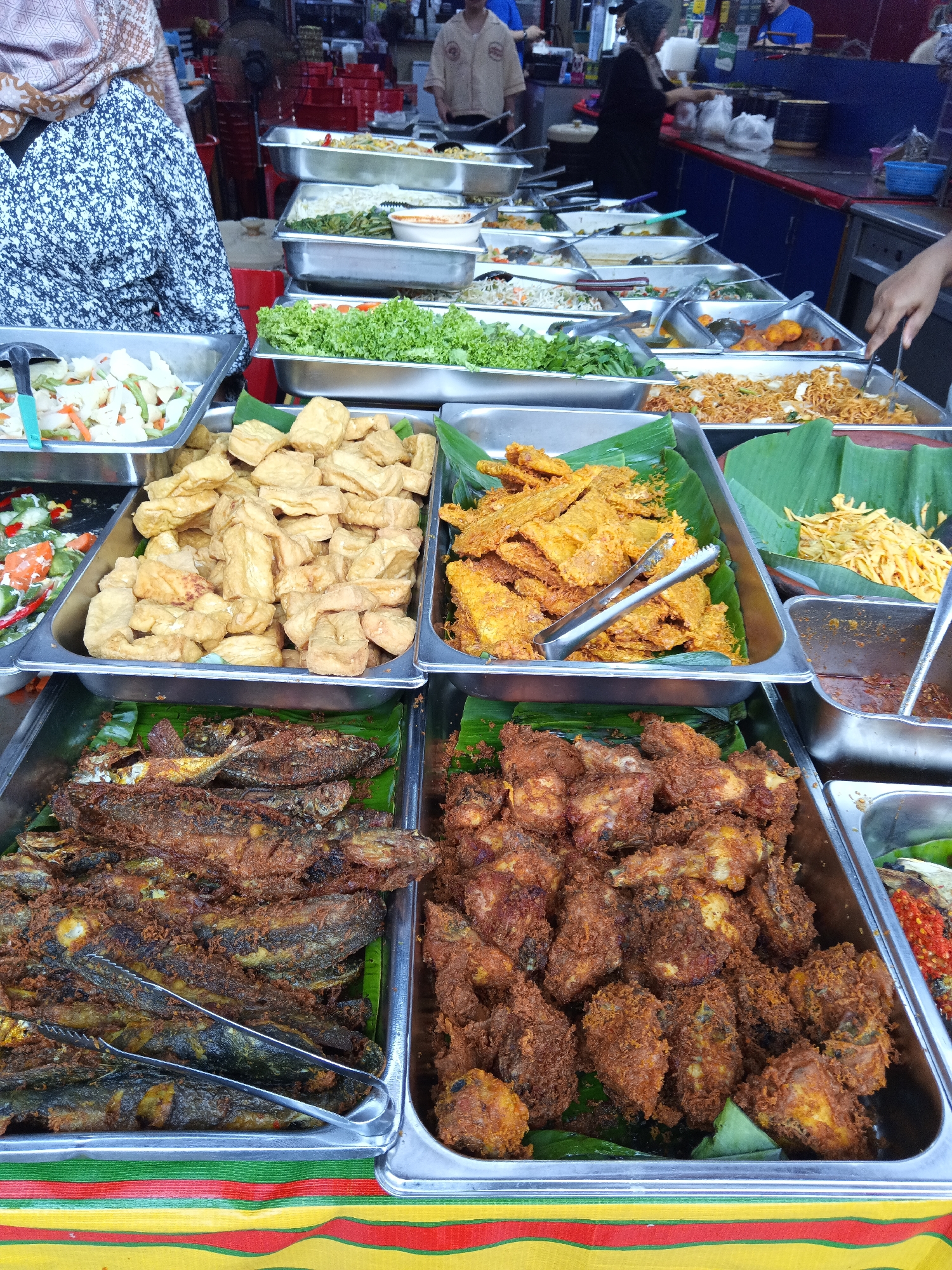 A street food stall in Kuala Lumpur, Malaysia displays a variety of prepared dishes. The foreground is dominated by several large metal trays filled with food. The tray on the far left contains an assortment of fried fish, laid out on banana leaves. To its right, another tray holds pieces of fried chicken, also on banana leaves. A third tray contains fried tofu cubes. Further back, other trays hold various dishes including fried noodles, vegetables, and what appears to be a spicy sauce. In the background, several people are visible, some standing behind the food stalls and others browsing the offerings. The stall appears to be set up outdoors, likely in a busy market or along a street. The overall impression is one of a vibrant food market, offering a diverse selection of local cuisine.