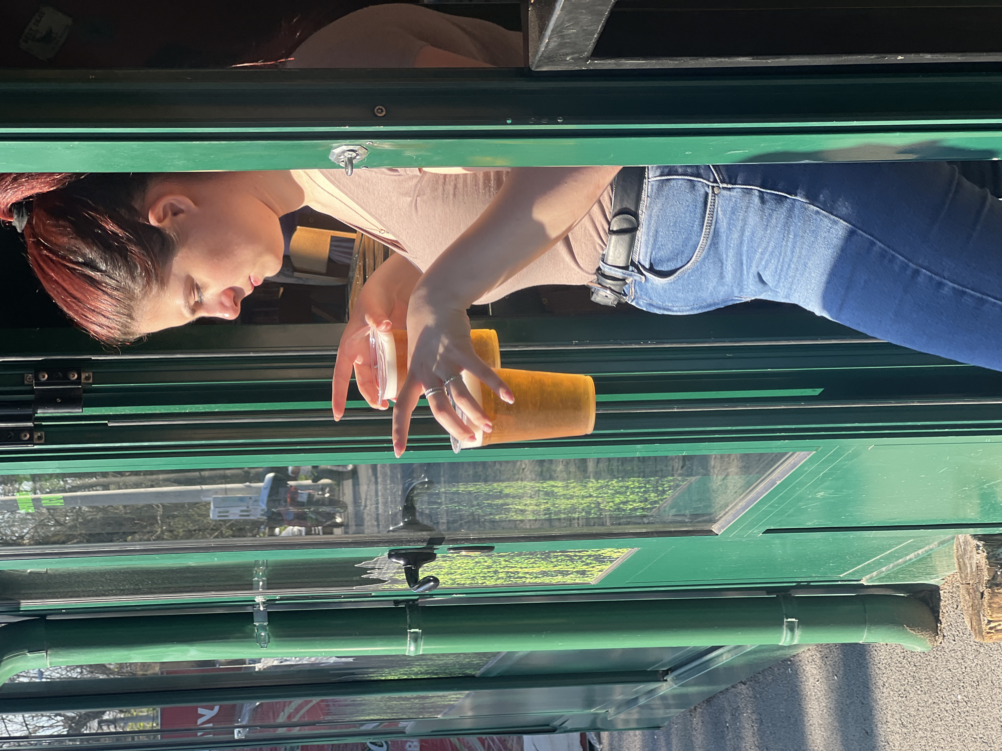 A young woman with red hair stands inside a green building. She wears blue jeans and a light-colored shirt. Her hands hold two translucent plastic cups filled with a yellow liquid. The scene appears to be a cafe or food service establishment. Through the window, a utility truck is visible. The location is Belgrade, Serbia.