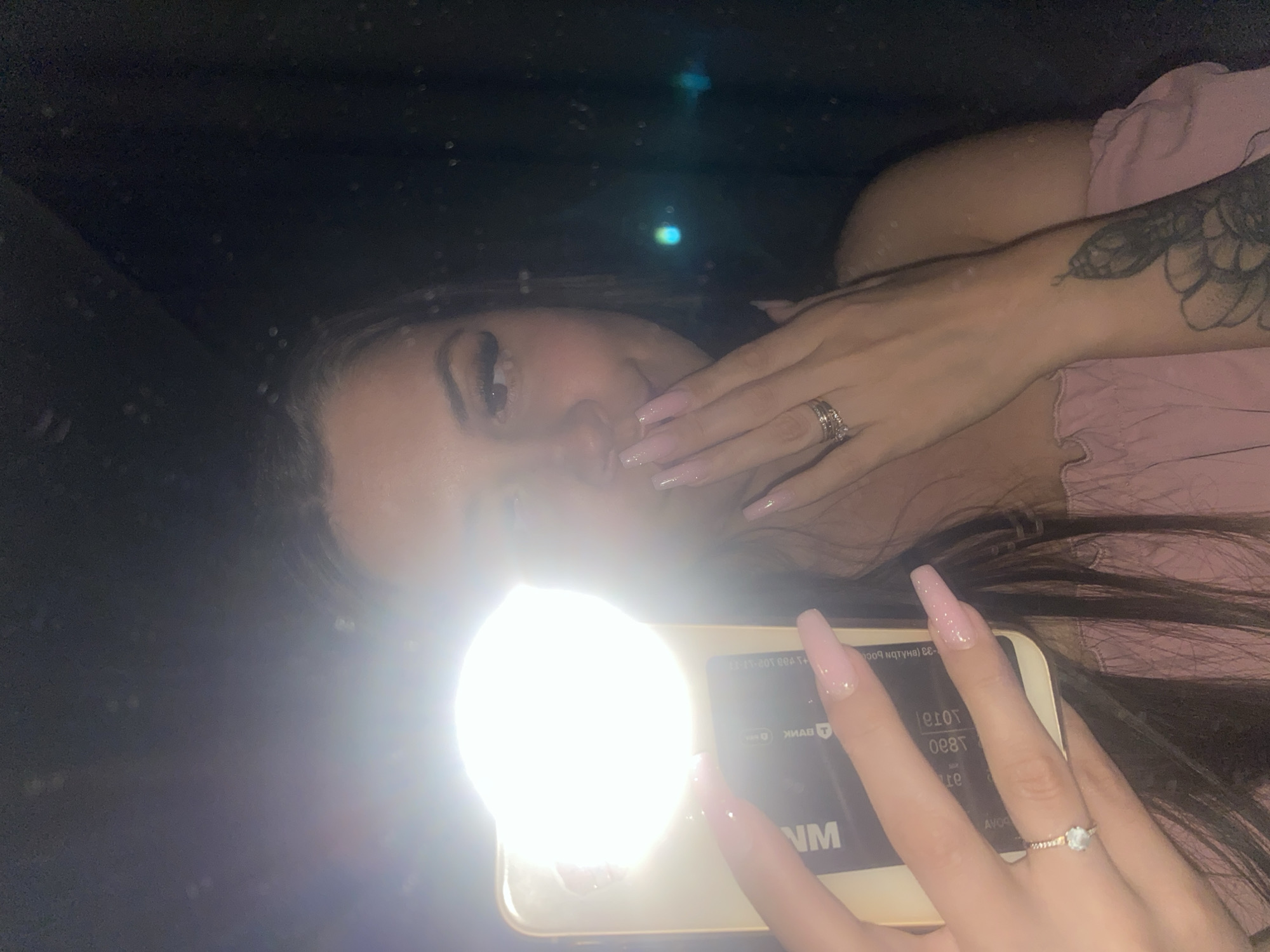 This is a selfie of a young woman taken at night. She is holding a smartphone with a bright ring light attached, which illuminates her face. She has long, light pink acrylic nails and is wearing a light pink off-the-shoulder top. She has on a ring with a small blue stone and another stacked ring. A tattoo of a flower and possibly a snake is visible on her left arm. The background is dark, suggesting an indoor or nighttime setting, possibly in a car, with some reflections visible. There is no information in the image to suggest it was taken in the city of Chaikovsky, Russia.