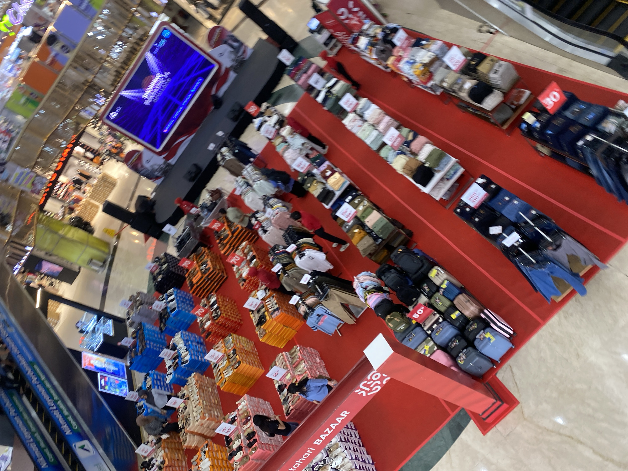 The image depicts an indoor bazaar, likely a department store, with a red carpeted floor and numerous display tables and stands. In the foreground, there are several rows of merchandise, including what appear to be folded clothing and bags. Further back, more items are displayed, some in blue crates and orange stacks, and there are also racks of clothing. Towards the upper right, a large screen is visible, displaying a promotional image. Several individuals are present, browsing the merchandise and interacting with displays, with some walking and others standing. The environment suggests a commercial setting, possibly a mall or large retail space, within the city of Banjarbaru, Indonesia.