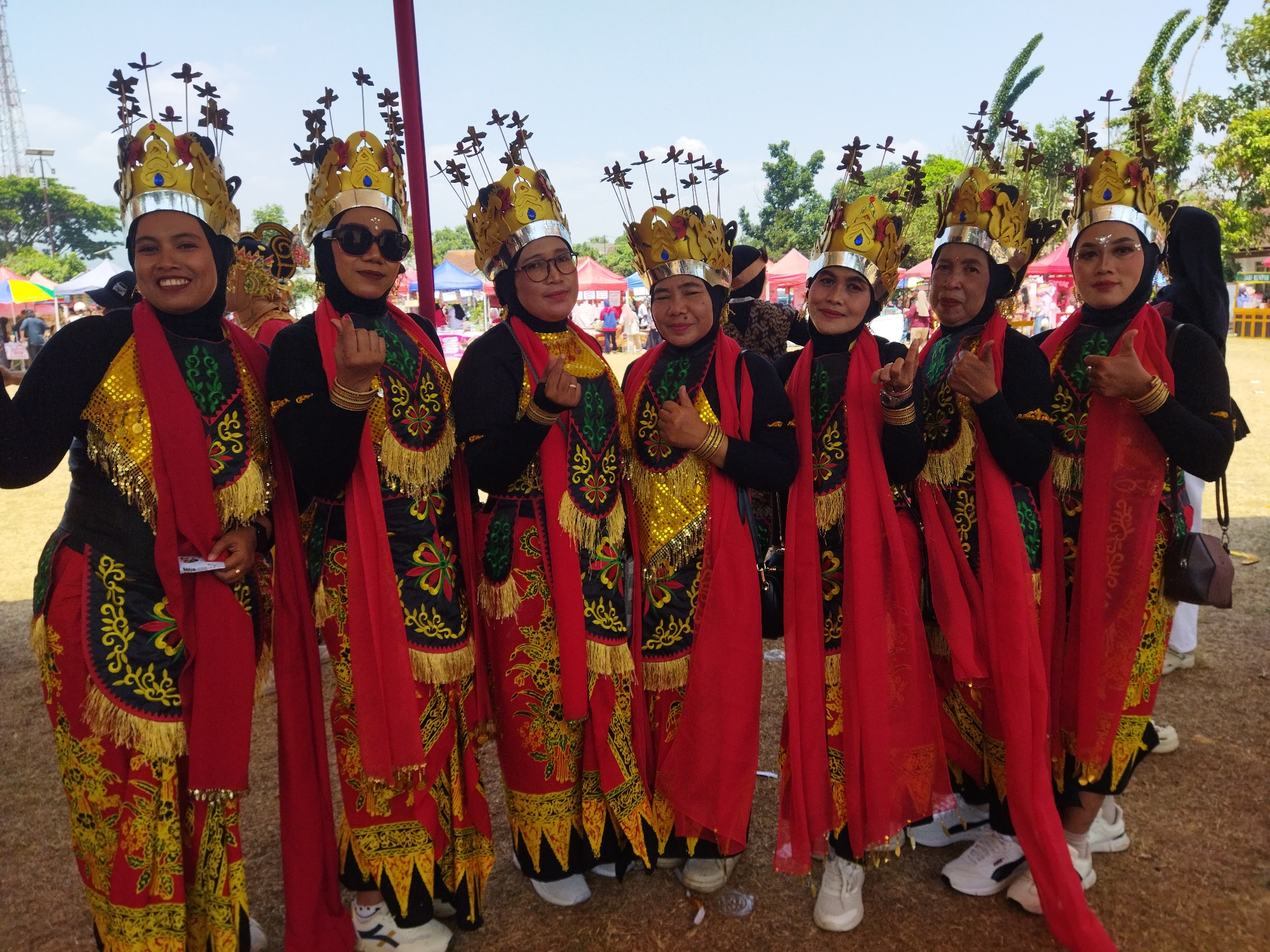 Seven women are posing for a photo in a group. They are all wearing traditional Indonesian attire, specifically the "kain tenun" fabric which is a type of woven textile, along with ornate headpieces and makeup. The women are dressed in black tops, vibrant red sashes, and colorful skirts with intricate patterns and gold embellishments. Their headpieces are elaborate, gold-colored crowns adorned with small, dark elements resembling leaves or feathers. They are standing outdoors on a dirt or gravel surface, possibly at a festival or cultural event. In the background, there are tents and trees, suggesting an outdoor venue. The sky is clear and blue, indicating a sunny day. The woman on the far left is smiling broadly and wearing dark lipstick. The woman second from the left is wearing sunglasses and making a hand gesture. The woman second from the right is giving a thumbs-up. The woman on the far right is also giving a thumbs-up. The location is the city of Semarang, Indonesia.