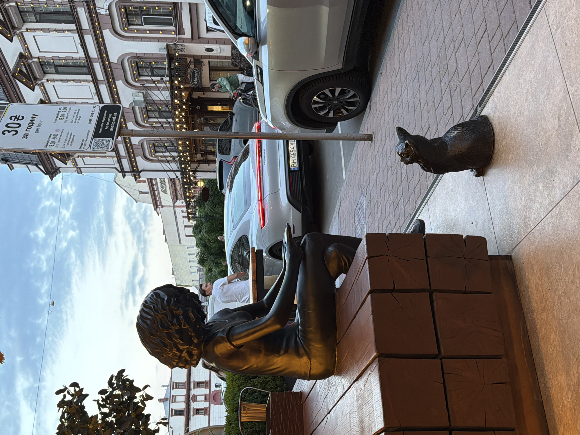 A bronze sculpture of a woman sitting on a wooden bench is positioned in the foreground of the image. The woman has curly hair and is holding a small object in her hands. To the right of the bench, a small bronze sculpture of a cat is seated on the pavement. In the background, there are parked cars, including a silver Porsche Taycan with license plate BH 4654 YE, and a gray Volvo SUV. A man on a bicycle is visible in the middle ground, near a building with a sign that reads "Mozart." A parking sign with pricing information is mounted on a pole in front of the building. The scene appears to be an outdoor seating area in a city, with buildings and street elements indicating an urban environment. The sky is cloudy.