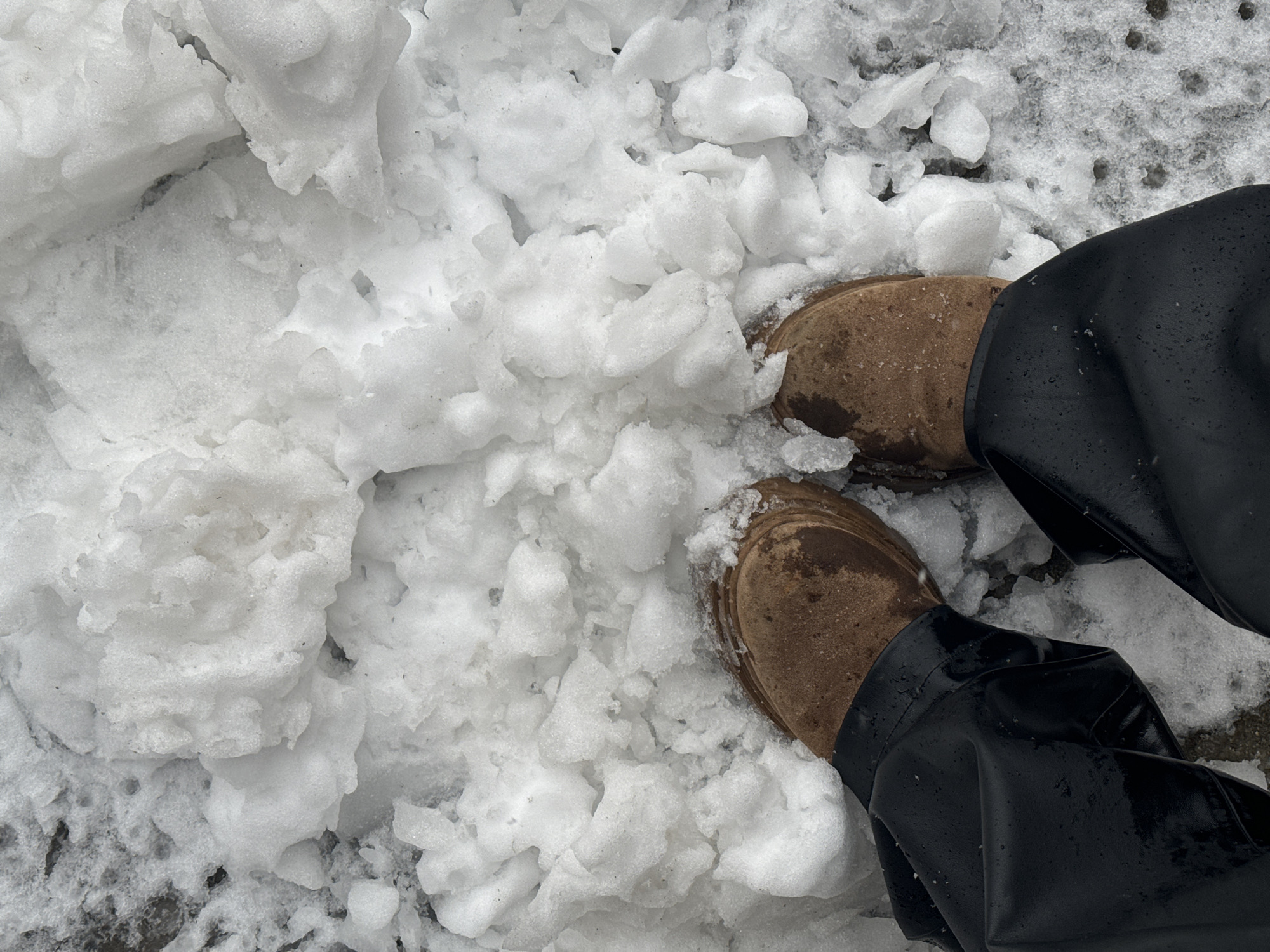 The image presents an overhead view of a person's lower legs and feet, partially submerged in white snow. The individual is wearing brown, possibly suede or leather, boots and dark, likely black, pants that appear to be made of a water-resistant material, as water droplets are visible on their surface.
The scene is set outdoors, characterized by significant snow accumulation on the ground. The snow varies in texture; some areas are fluffy and fresh-looking, while others are more compact, clumpy, or partially melted, revealing patches of darker ground beneath. The ground appears to be a paved or solid surface. The boots have some snow and possibly dirt on them, consistent with walking in snowy conditions. The overall impression is one of a cold, wintery environment.
There is no discernible activity beyond standing or pausing in the snow. No specific time of day is indicated, though it is clearly daylight. No text is visible in the image. The location is specified as Odintsovo, Russia.