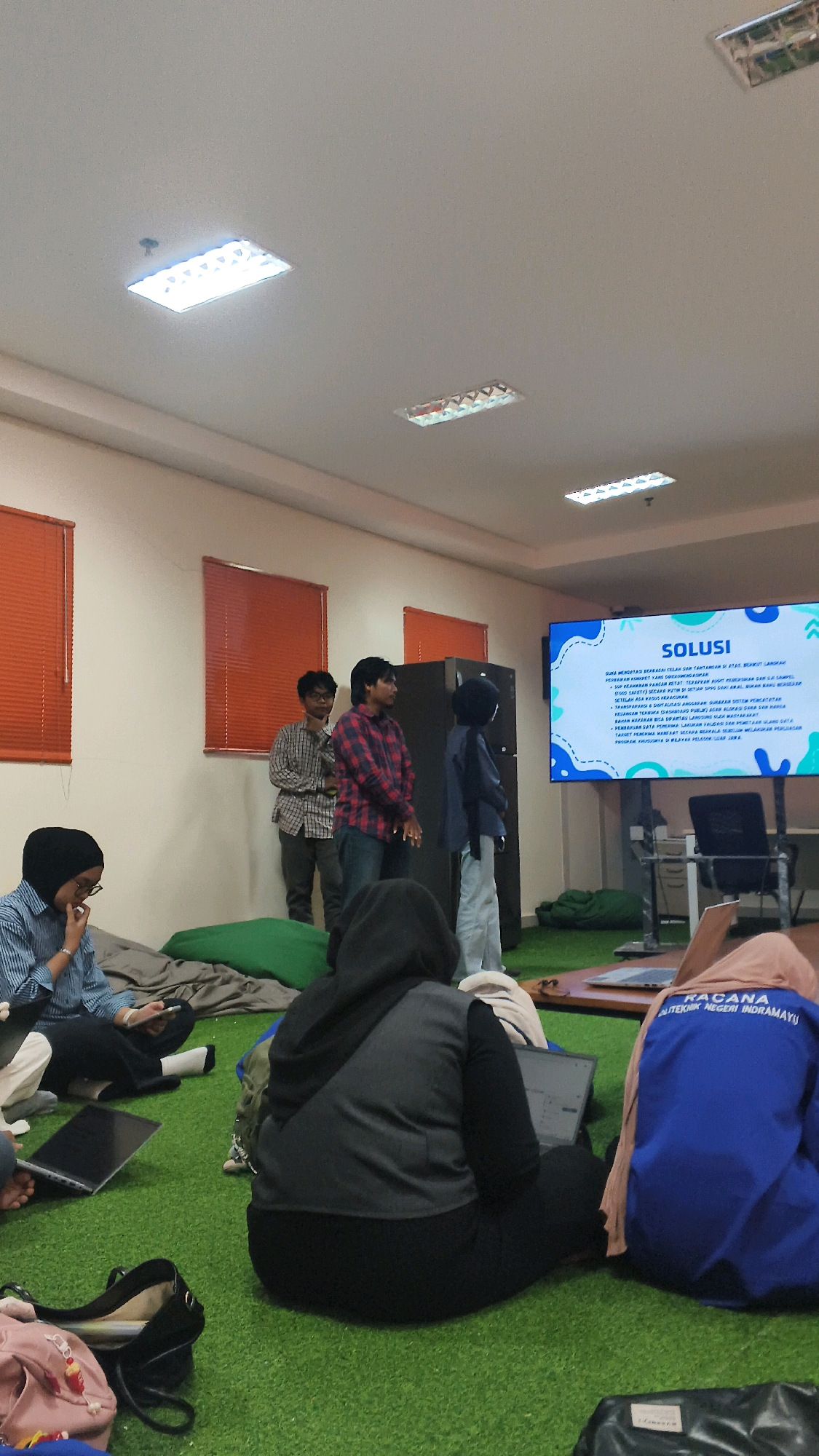 The image depicts an indoor gathering in a room with artificial turf flooring, suggestive of a casual or creative workspace. The room is lit by overhead fluorescent lights. Several individuals are present, with most seated on the floor, some engaging with laptops. The focal point of the scene is a large digital display screen showing a presentation with the heading "SOLUSI" (Solution in Indonesian). To the right of the screen is a filing cabinet and some office furniture. To the left, three men stand, appearing to be presenting or discussing the material on the screen. The room has windows with orange blinds. Based on the visible text on the screen and the clothing of the attendees, it is highly probable that this scene is located within an educational institution or a professional setting in Lohbener, Indonesia, possibly related to a presentation or workshop.