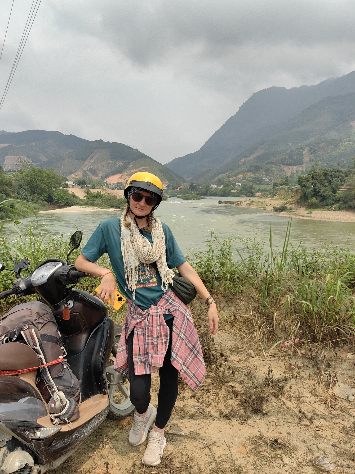 A lone individual stands roadside in Lang Qua, Vietnam, leaning against a scooter. The setting features a wide, pale green river flowing from left to right across the midground, flanked by steep, forested mountains under a cloudy, overcast sky. The mountains exhibit patches of exposed reddish-brown soil, indicating erosion or deforestation. Scattered small buildings are visible on the far bank of the river.
The individual, a female, stands facing forward, slightly turned to her left, with her right arm resting on the handlebars of a dark-colored scooter. She wears a yellow open-face helmet with black sunglasses, a light-colored patterned scarf, a dark teal t-shirt, black leggings, and light athletic shoes. A red and white plaid shirt is tied around her waist. She holds a bright yellow smartphone in her right hand and has a black fanny pack or sling bag across her chest. Her left wrist displays multiple dark bracelets, and her right wrist has a beaded orange bracelet. The scooter has a dark brown backpack secured to its front floorboard area with a red strap, and keys are in the ignition. A phone mount is attached to the handlebars. The immediate foreground consists of a dirt and sparse grass ground with some small white flowering plants on the riverbank to the left. Overhead, power lines are visible in the upper left quadrant of the frame. Debris, including a plastic bottle and a sandal, lies on the dirt ground to the right.
