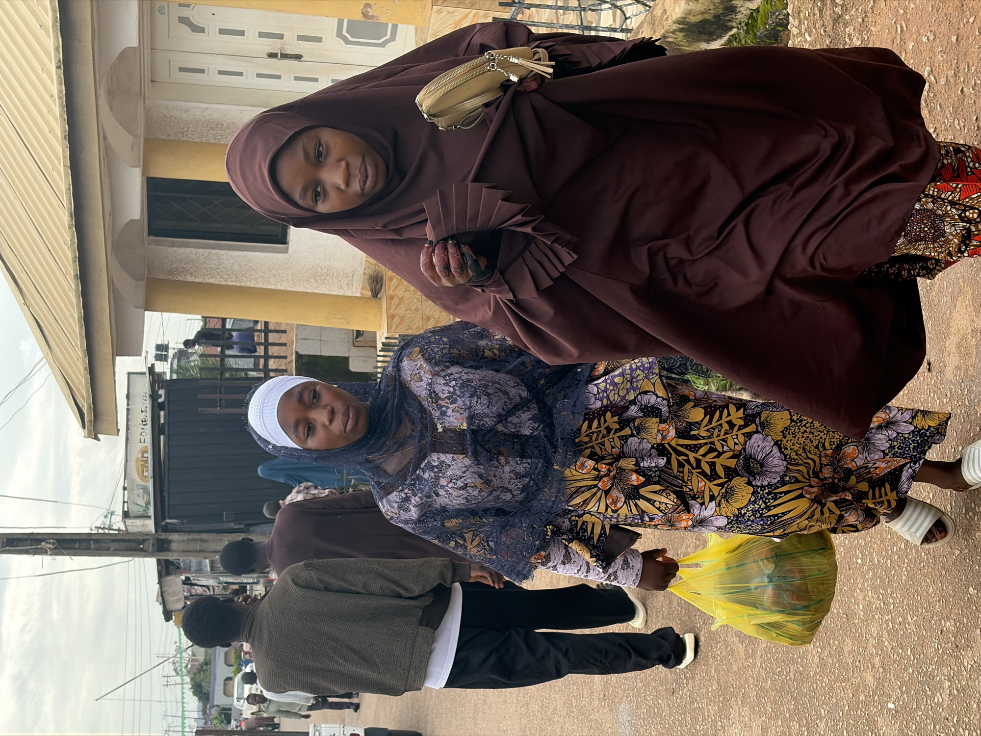 Two women, likely Nigerian and dressed in modest Islamic attire, walk along a dusty, unpaved road in Jos, Nigeria. The woman on the left wears a light blue hijab and a long, colorful dress with a floral pattern. She carries a bright yellow plastic bag filled with produce. The woman on the right is cloaked in a dark brown hijab and dress, and she clutches a small, tan purse. Behind them, other pedestrians and vehicles are visible, suggesting a bustling street scene. The architecture in the background features simple, concrete buildings with corrugated metal roofs, typical of many areas in Jos. The sky is overcast, indicating a cloudy day.