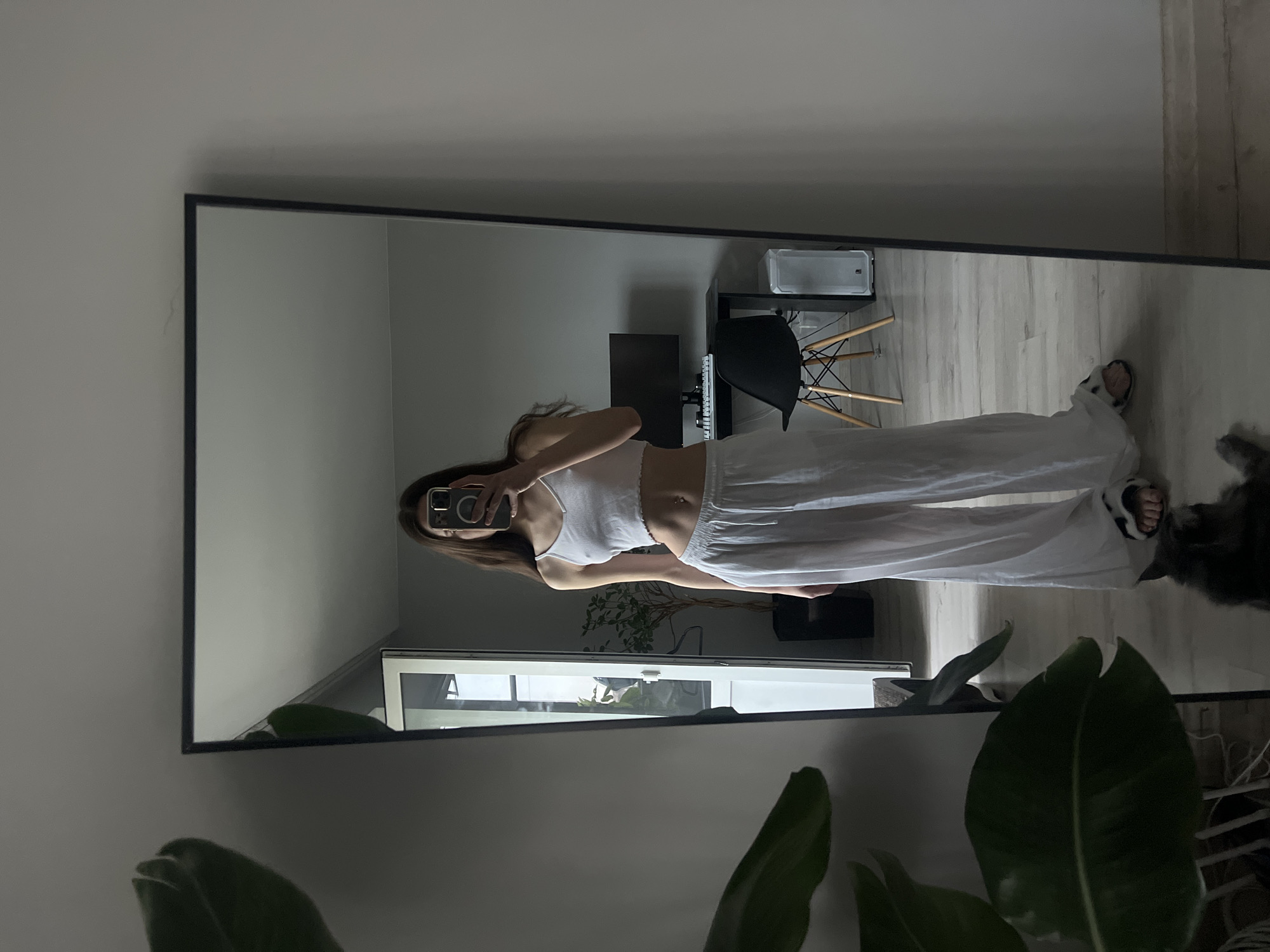 A full-length mirror reflects a woman in a white crop top and white pants, taking a photo of herself with a smartphone. A desk and chair are visible in the background. A potted plant sits at the bottom of the frame. Location is not provided, thus it cannot be included in the description.