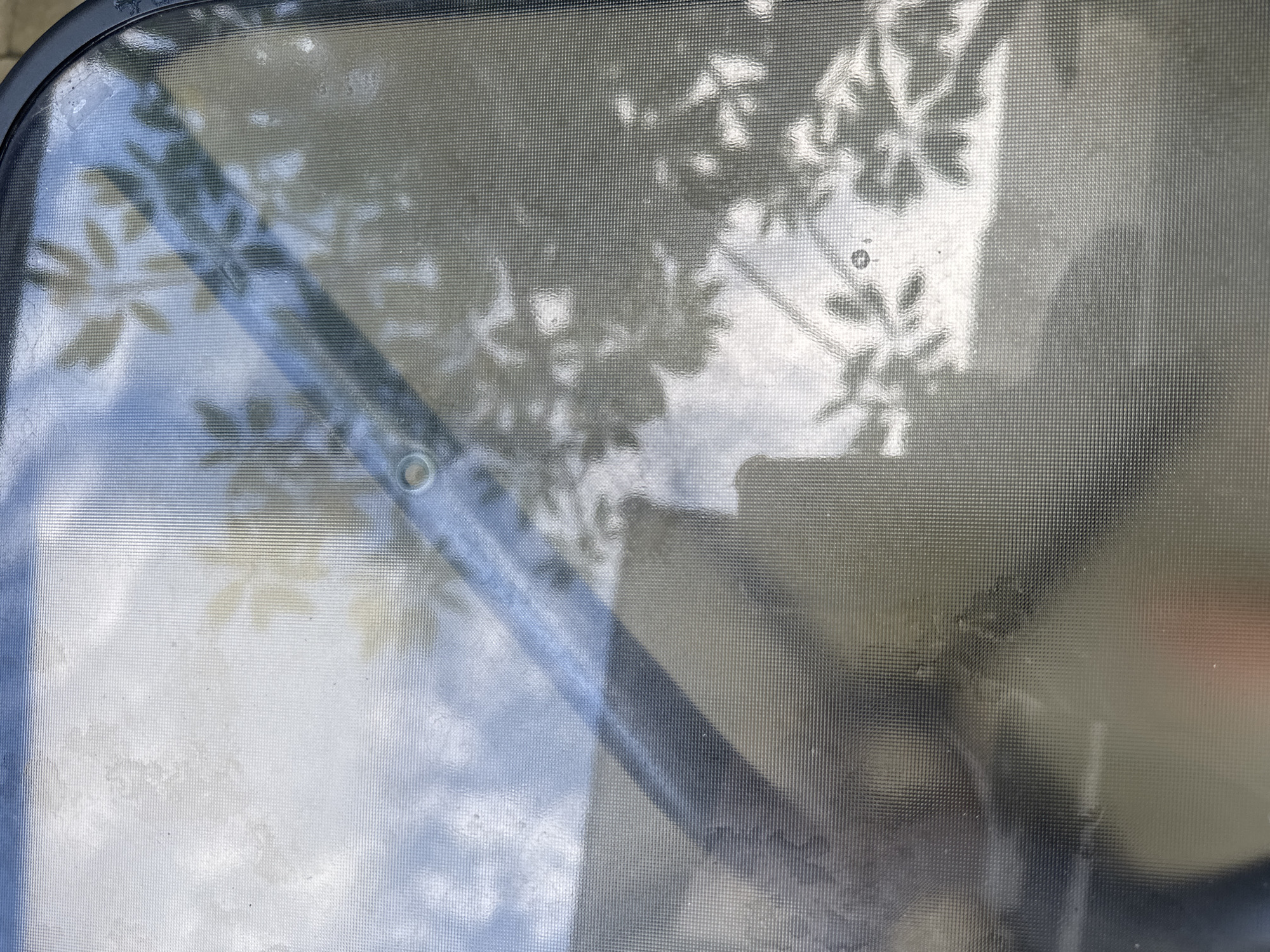 The image shows a close-up view through a textured glass or plastic surface, possibly a window or cover. There are reflections of foliage, suggesting a tree or bush is outside. A dark, linear object, possibly a frame or structural element, runs diagonally across the scene. Light and shadow play across the textured surface, creating a distorted view of the outside environment. The lack of further context prevents a specific determination of the scene or its relation to Kuta Selatan, Indonesia.