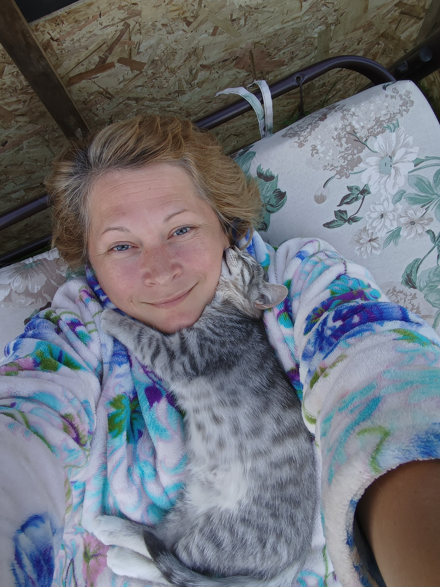 An overhead, medium shot captures a woman with short blonde hair and blue eyes, her face slightly angled towards the camera, a gentle smile on her lips. She is wearing a plush, colorful bathrobe with a pattern of white, blue, green, and purple swirls. Cuddled against her neck and shoulder is a small, grey tabby kitten, its eyes closed in apparent contentment. The woman's arm is visible, draped over the kitten, and her left hand is resting near her face. They are situated on a floral-patterned cushion, part of a swing seat. Behind them, rough, particleboard-like material forms a canopy, with a dark metal frame visible above. The scene suggests a relaxed moment, likely outdoors, given the natural lighting and the outdoor furniture. This image was taken in Ivanovskoe, Russia.