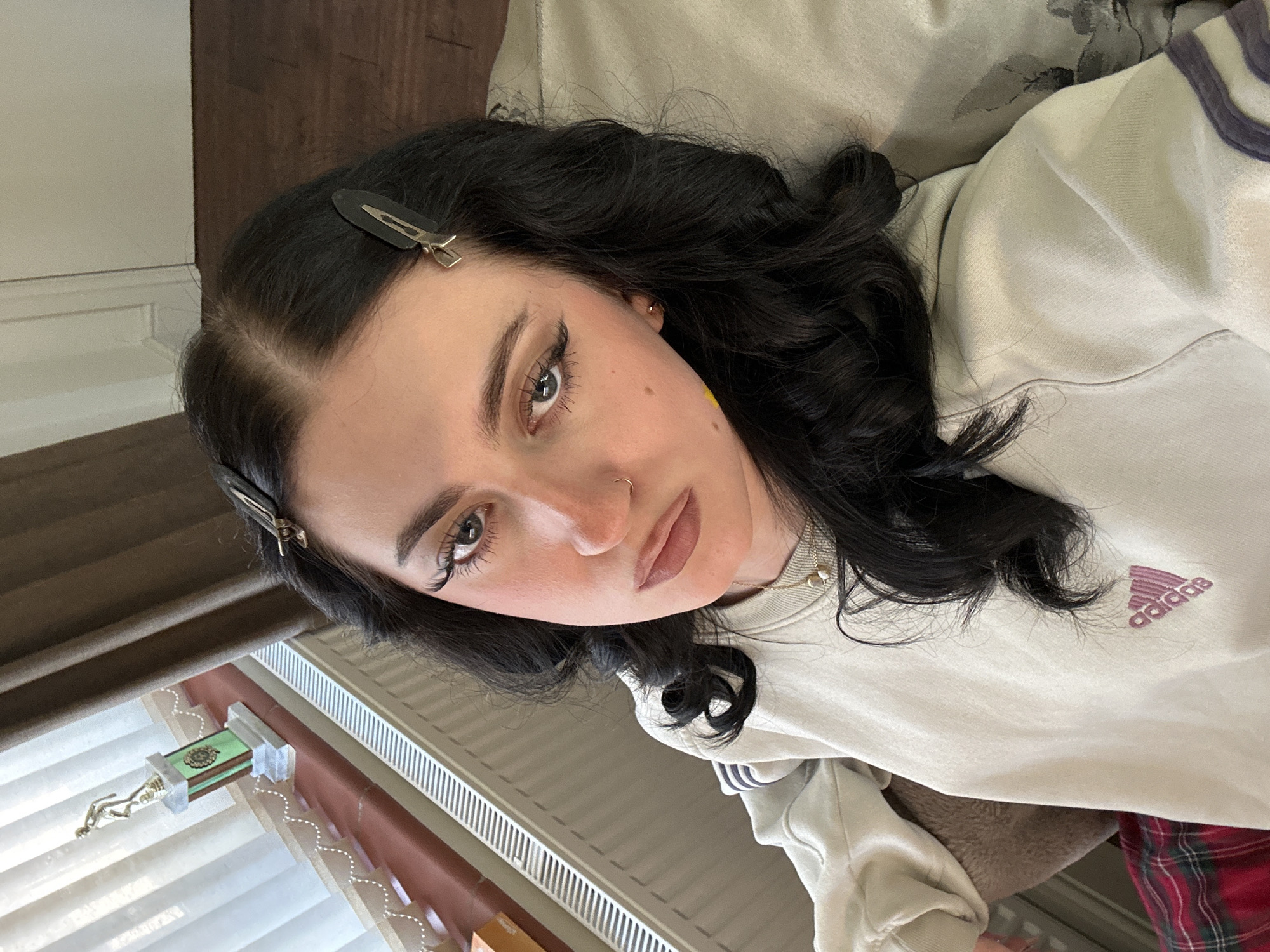 A medium close-up shot from a high angle captures a young woman with dark hair and fair skin. She is wearing an off-white Adidas sweatshirt. Her hair is styled with clips and loose curls. She has fair eyebrows, dark eyeliner, and a nose ring. The background shows a window with white blinds, a brown curtain, and a trophy on the windowsill. The image also includes a glimpse of a wooden floor and a patterned pillow or cushion. There is no specific identifiable information in the photo to precisely place the location in North Hertfordshire. The setting appears to be indoors, possibly in a residential dwelling.