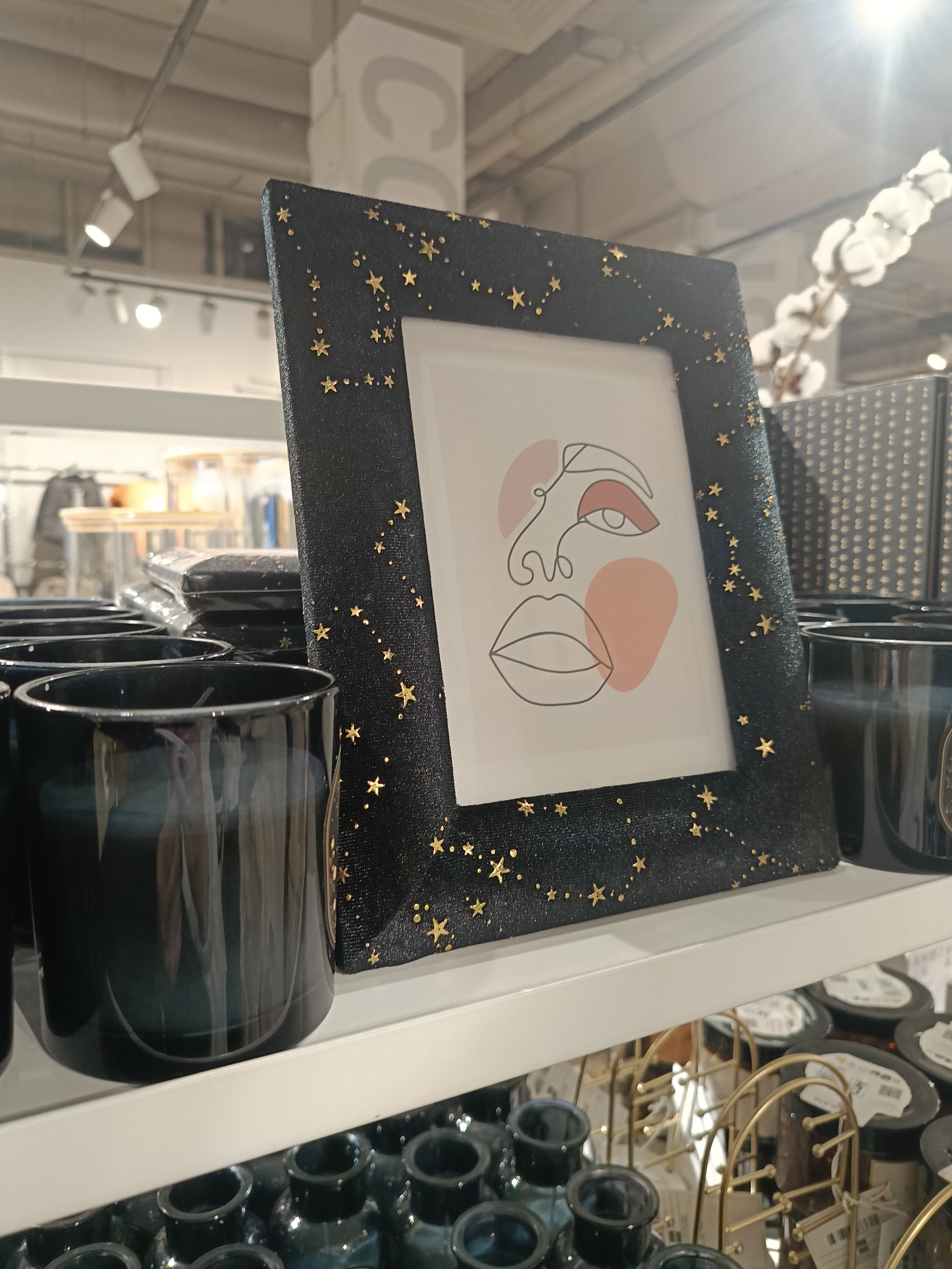 A slightly tilted medium shot taken indoors shows a black framed picture on a shelf in Izhevsk, Russia. The picture features a white background with a stylized drawing of a face in black line art with some sections filled in with light red. The frame is black with small gold star decorations. To the left of the picture are several shiny black cylindrical objects, possibly vases or candle holders. Beneath the shelf are numerous small blue-green glass vases and gold-colored metal racks with unidentified objects. In the background are partially obscured objects, suggesting a retail setting. The lighting is bright, with a visible spotlight in the upper right corner of the frame.